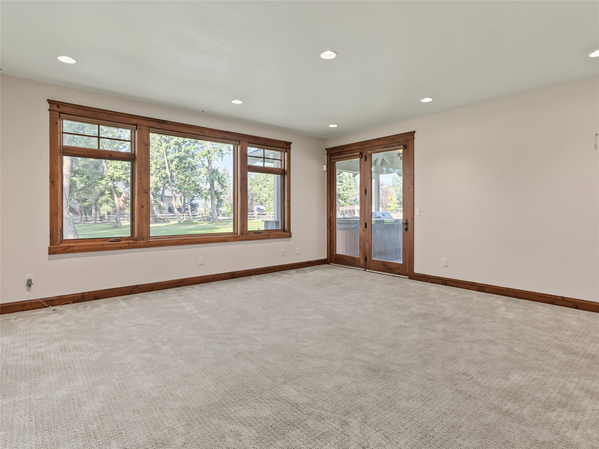 Empty carpeted living area with broad wood-trimmed windows and glass doors facing a tree-lined lawn.