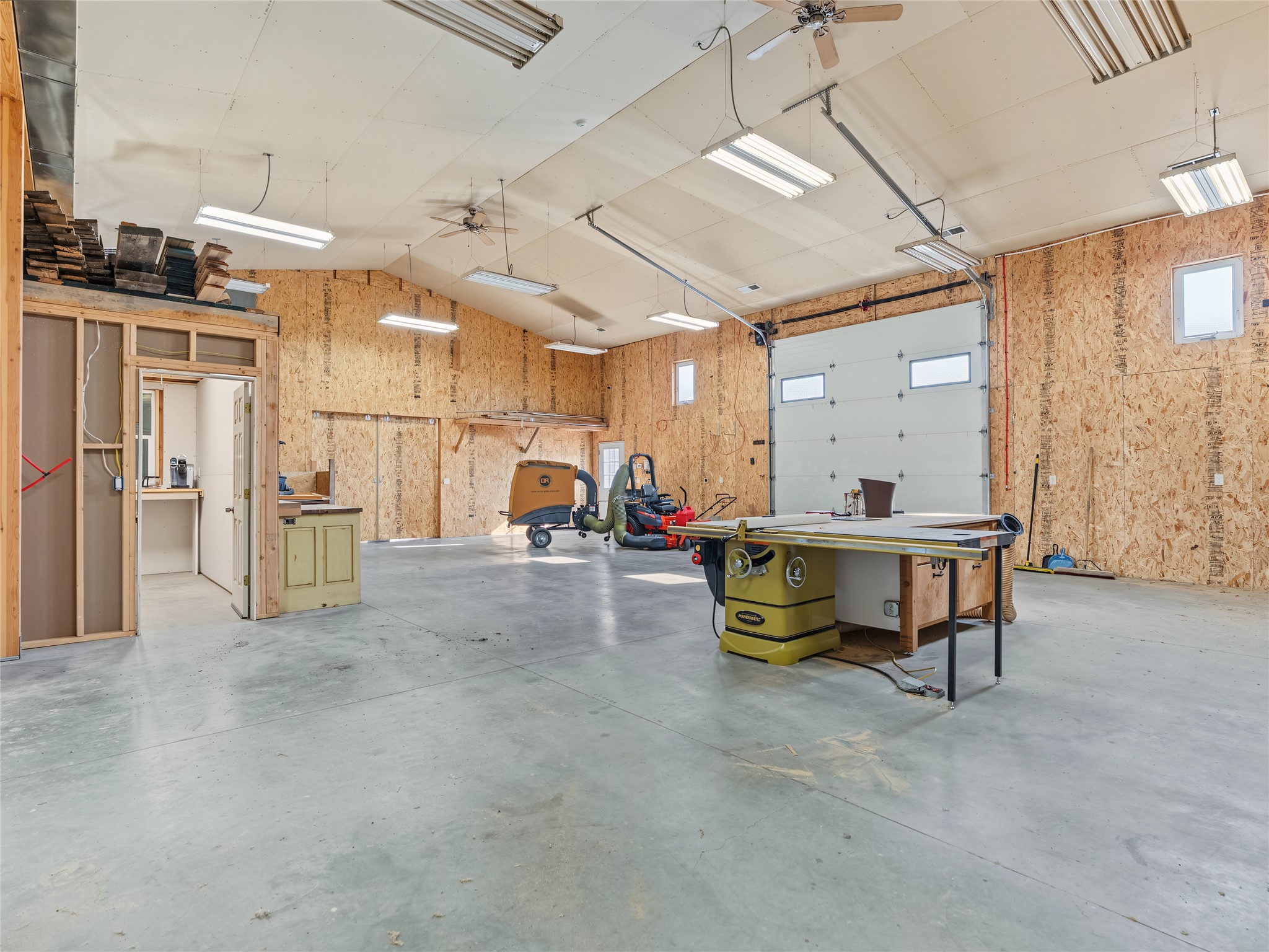 Spacious woodworking workshop with a table saw, exposed OSB walls, and a large garage door.