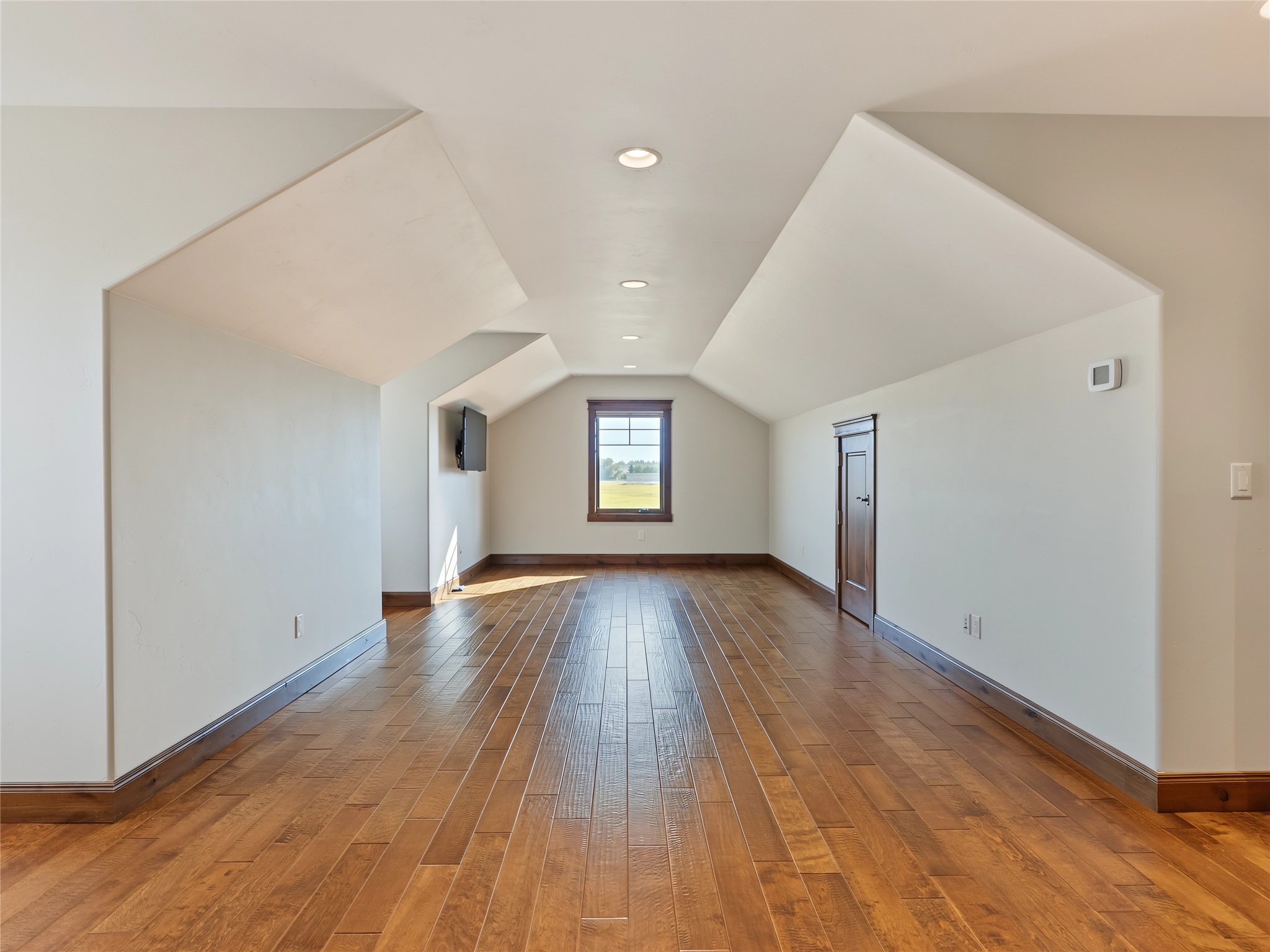 Empty vaulted room with warm wood flooring, a far window, and a wall-mounted television.