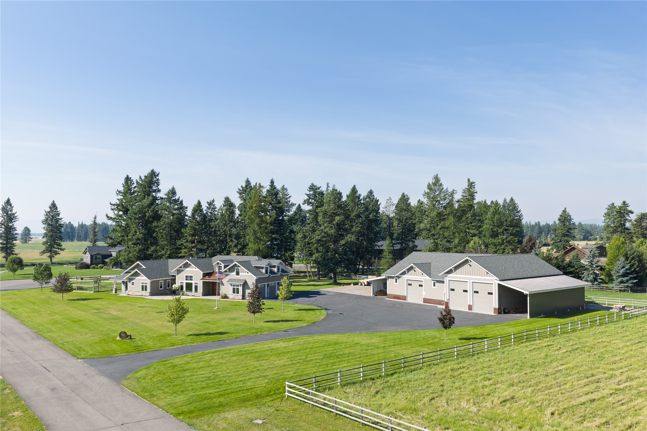 Rural home and large detached workshop surrounded by lawns, evergreen trees, and white rail fencing.