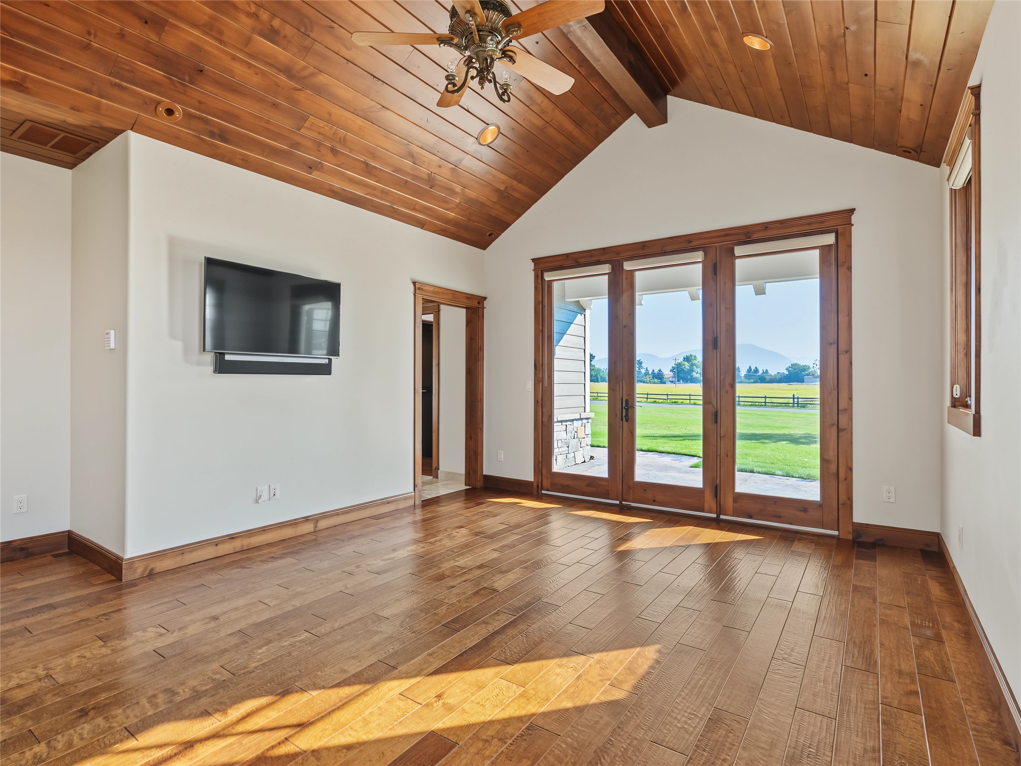 Vacant living room with vaulted wood ceiling, hardwood floors, and glass doors overlooking lawn and mountains.