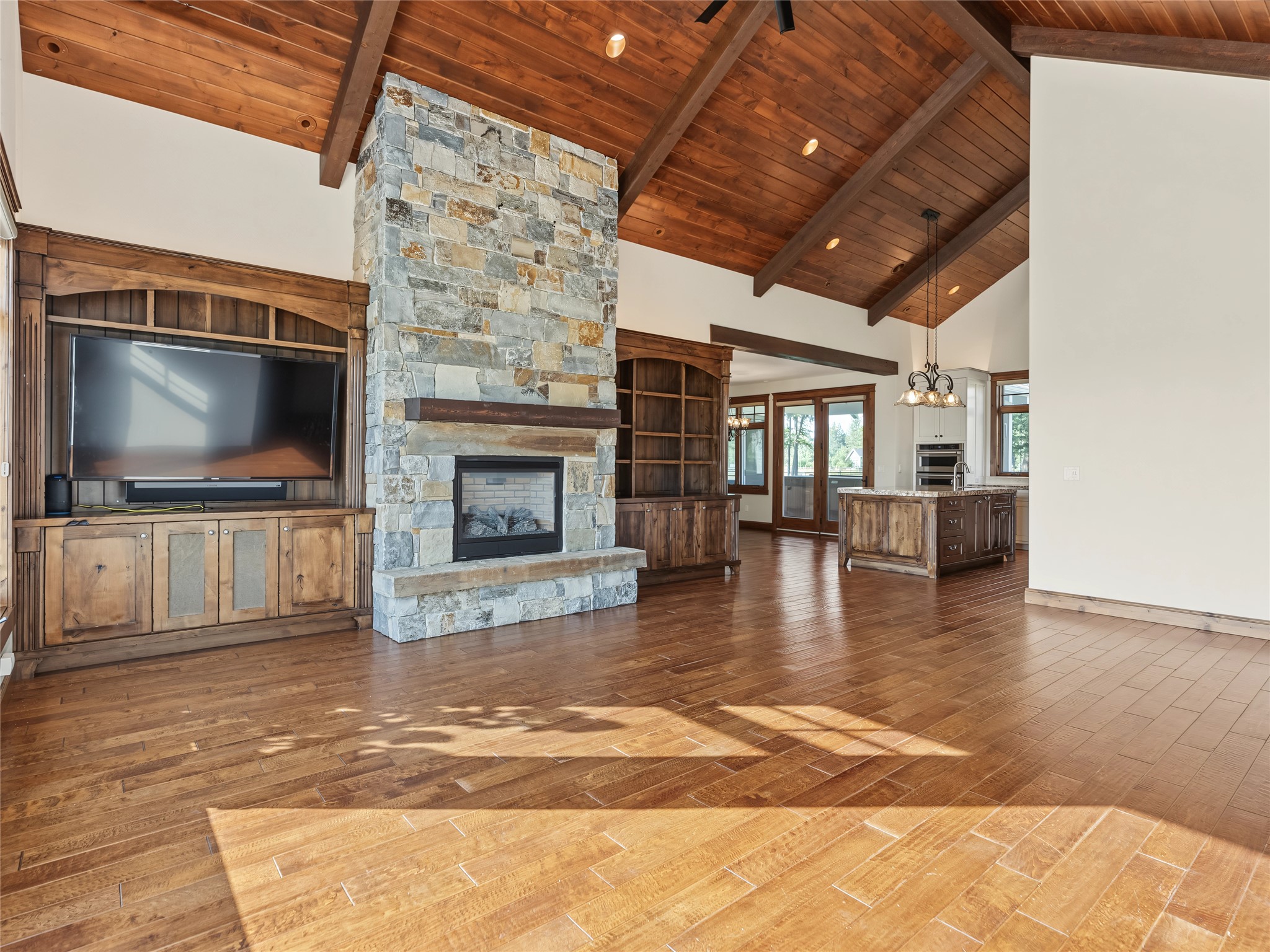 Living room with a tall stone fireplace, built-in television cabinetry, vaulted wood ceiling, and adjoining kitchen island.