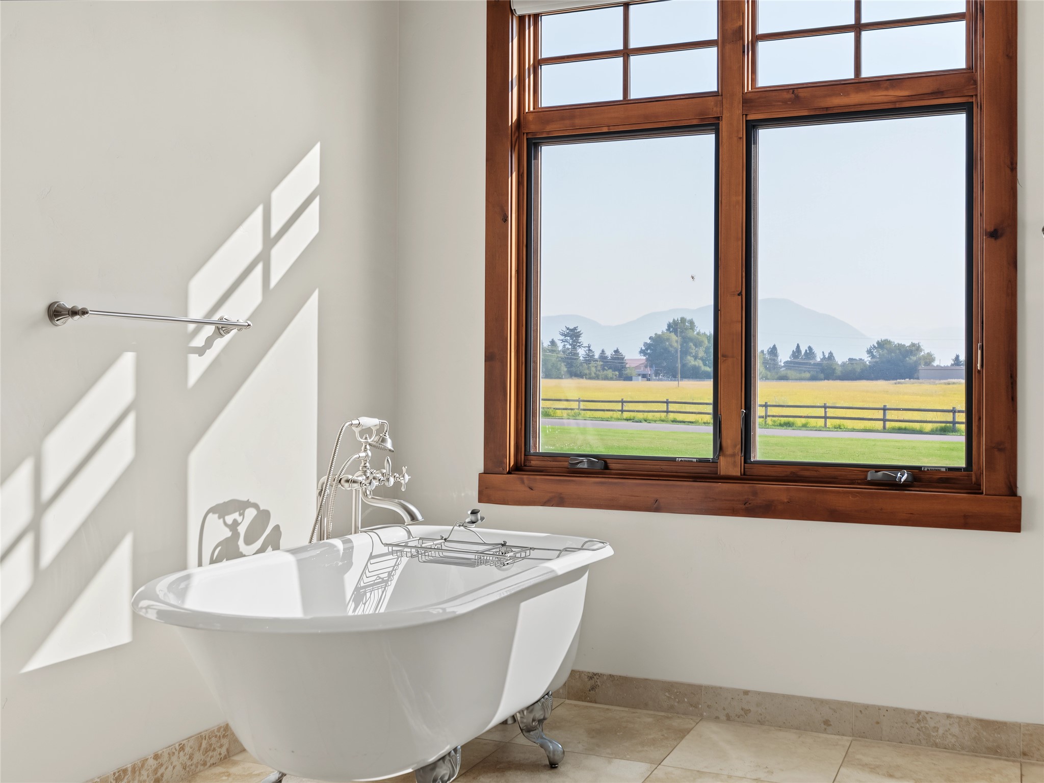 Freestanding clawfoot tub beneath large windows overlooking a fenced field and distant mountains.