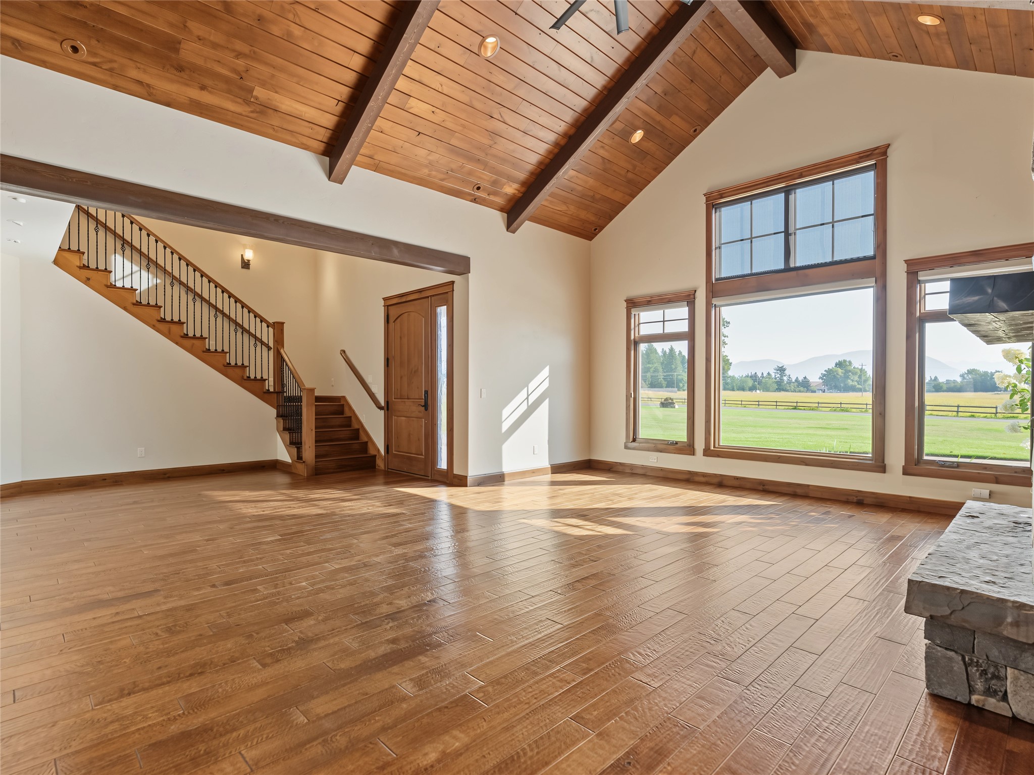 Vacant living room with vaulted wood ceiling, hardwood floors, staircase, and mountain views through large windows.