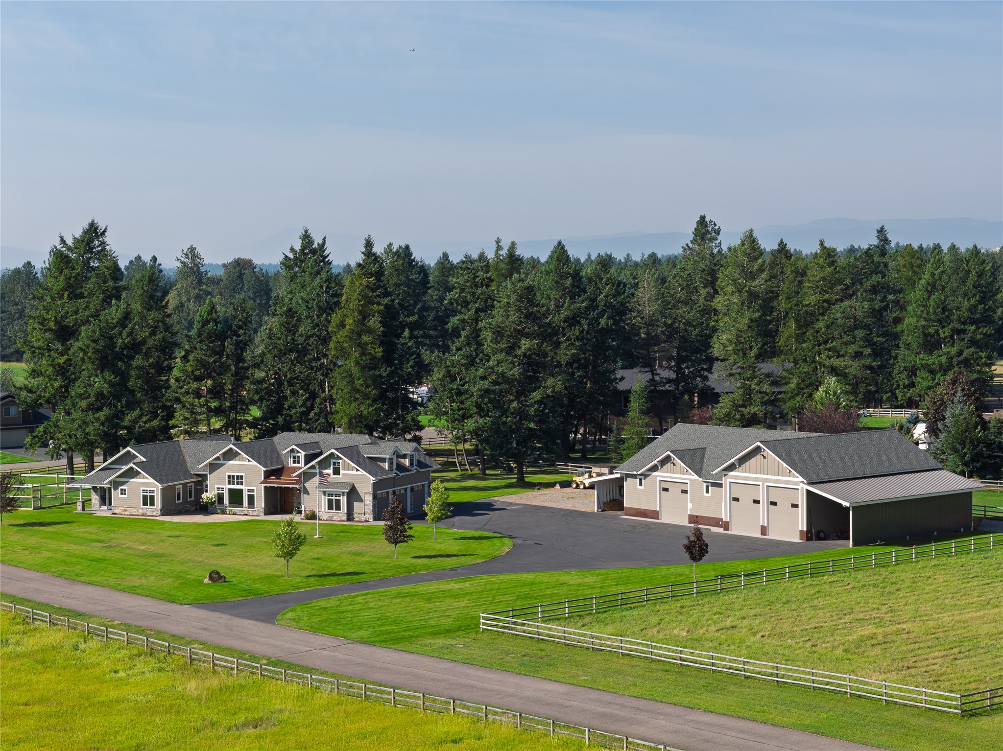 Rural home and detached multi-bay garage amid fenced lawns, evergreen trees, and distant mountains.