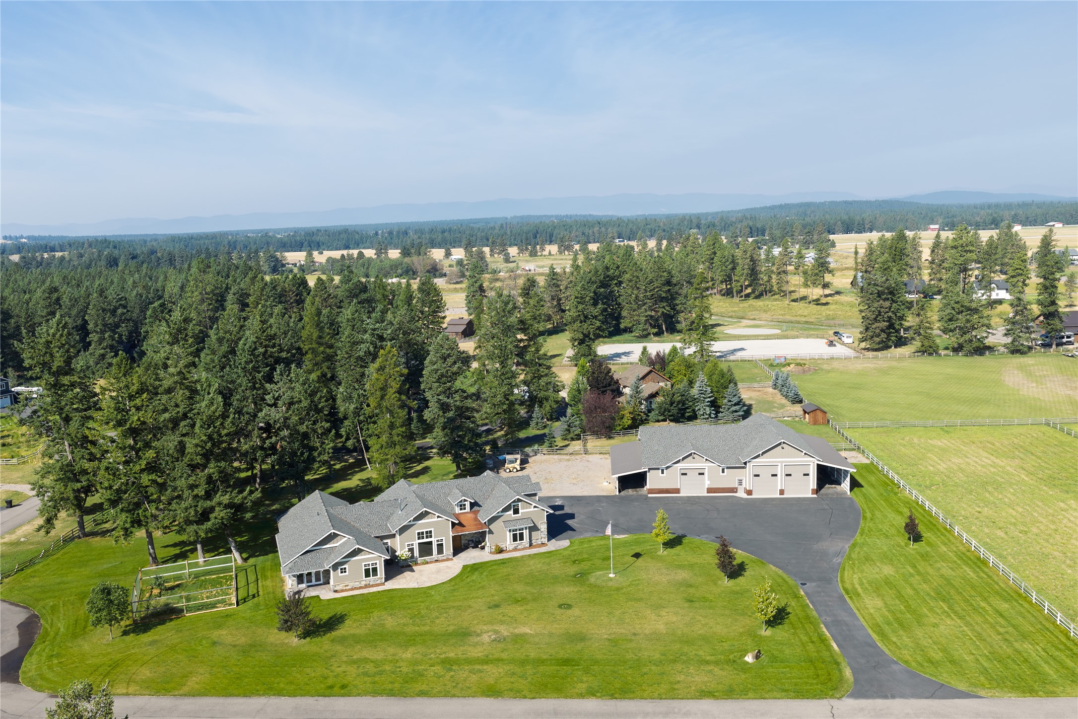 Aerial view of a gray rural home and large detached garage amid lawns, fenced fields, and evergreen trees.