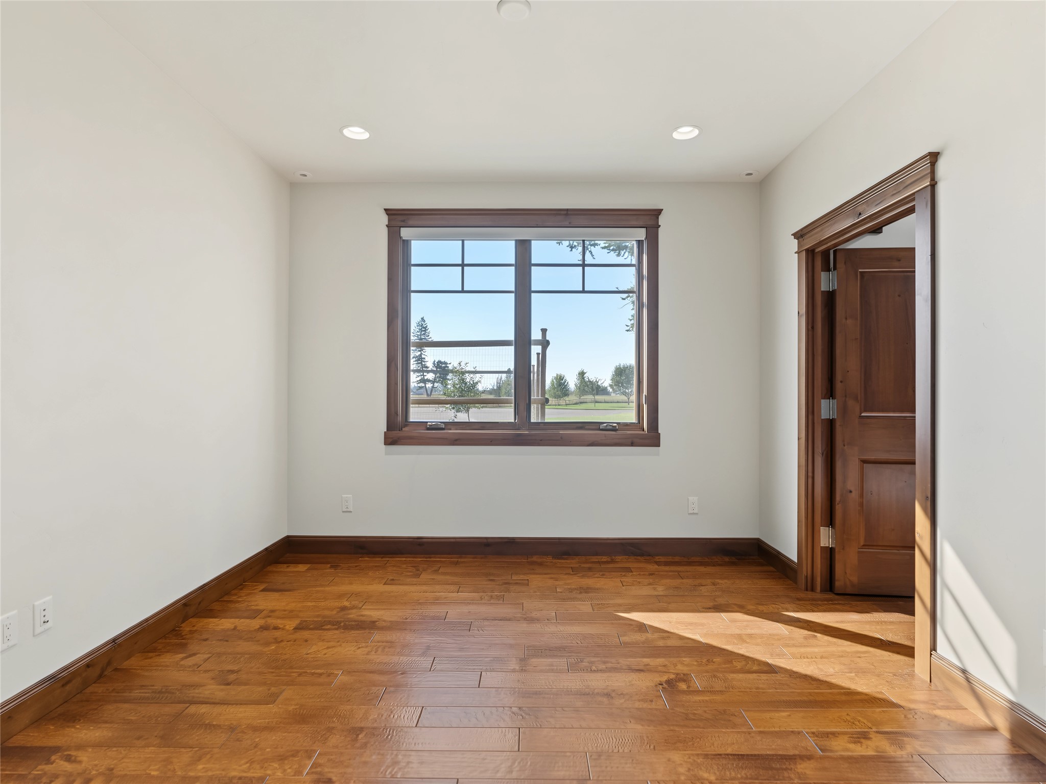 Empty room with warm wood flooring, dark wood trim, and a large window overlooking a lawn.