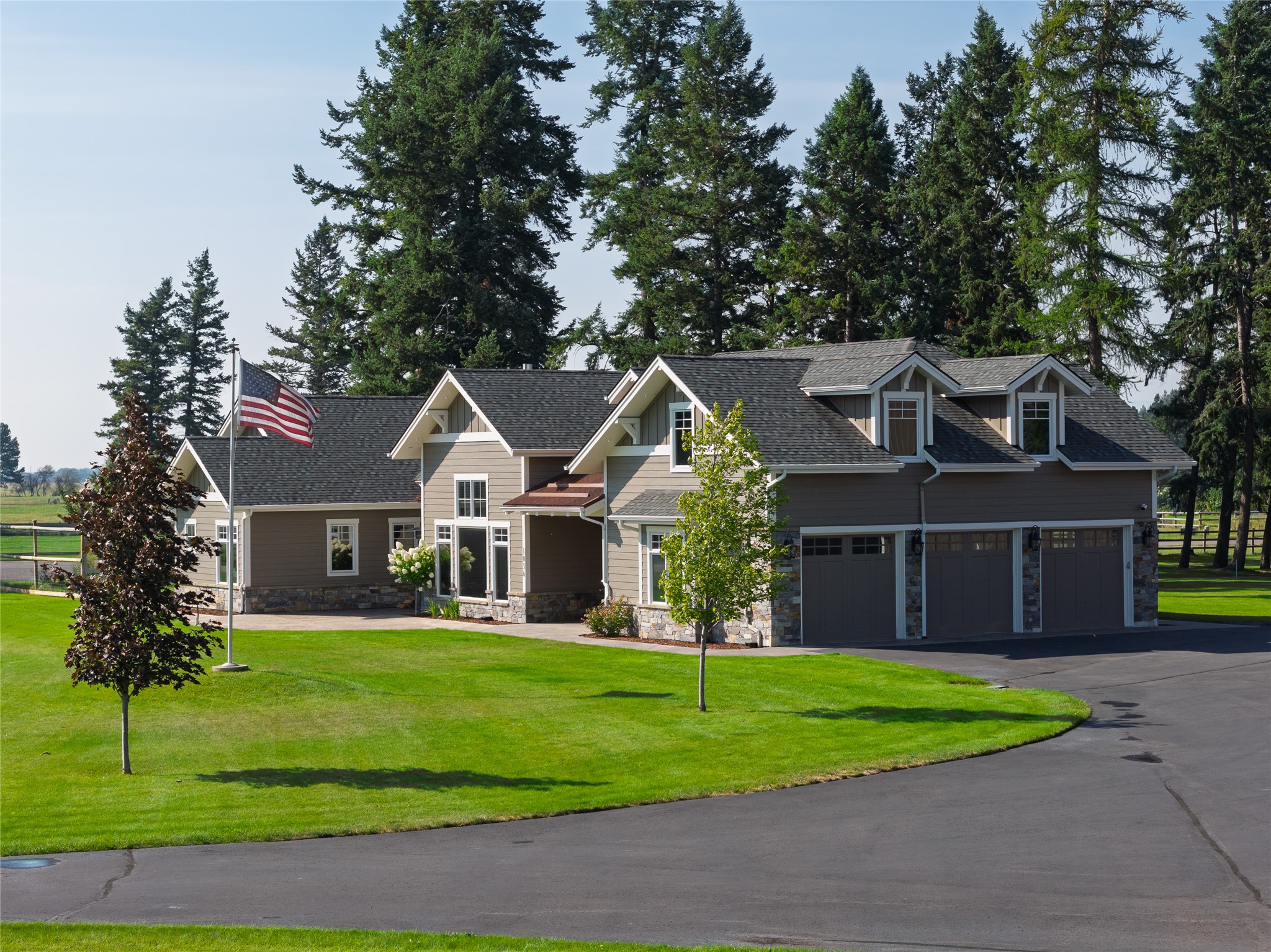 Large gray-brown home with a three-car garage, green lawn, mature evergreens, and an American flag.