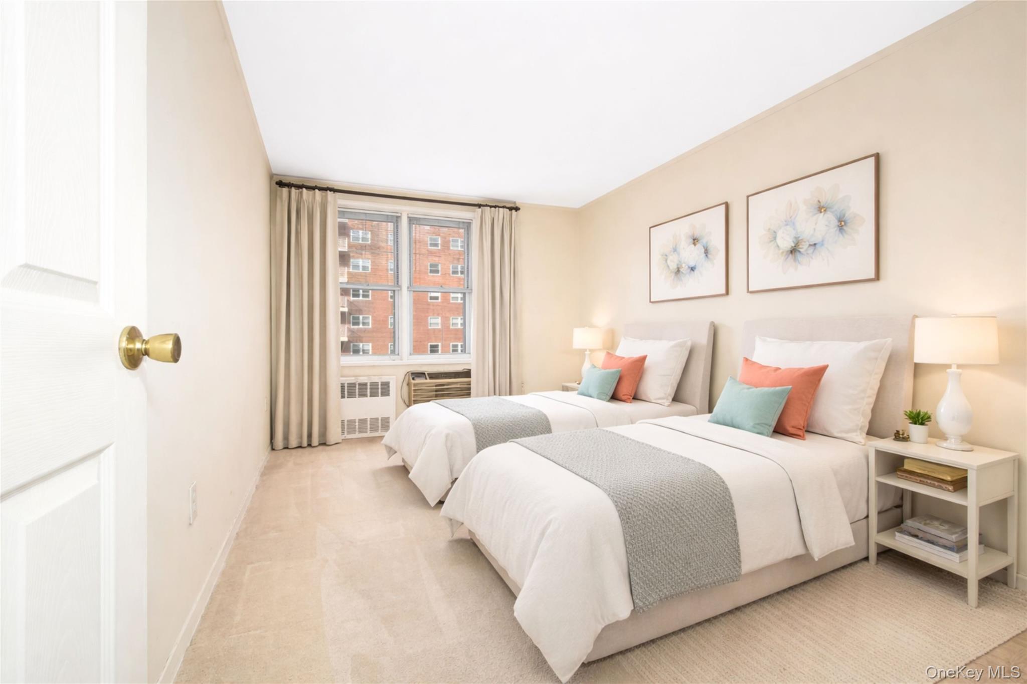 Staged bedroom with two beds, colorful accent pillows, and a window overlooking a brick building.
