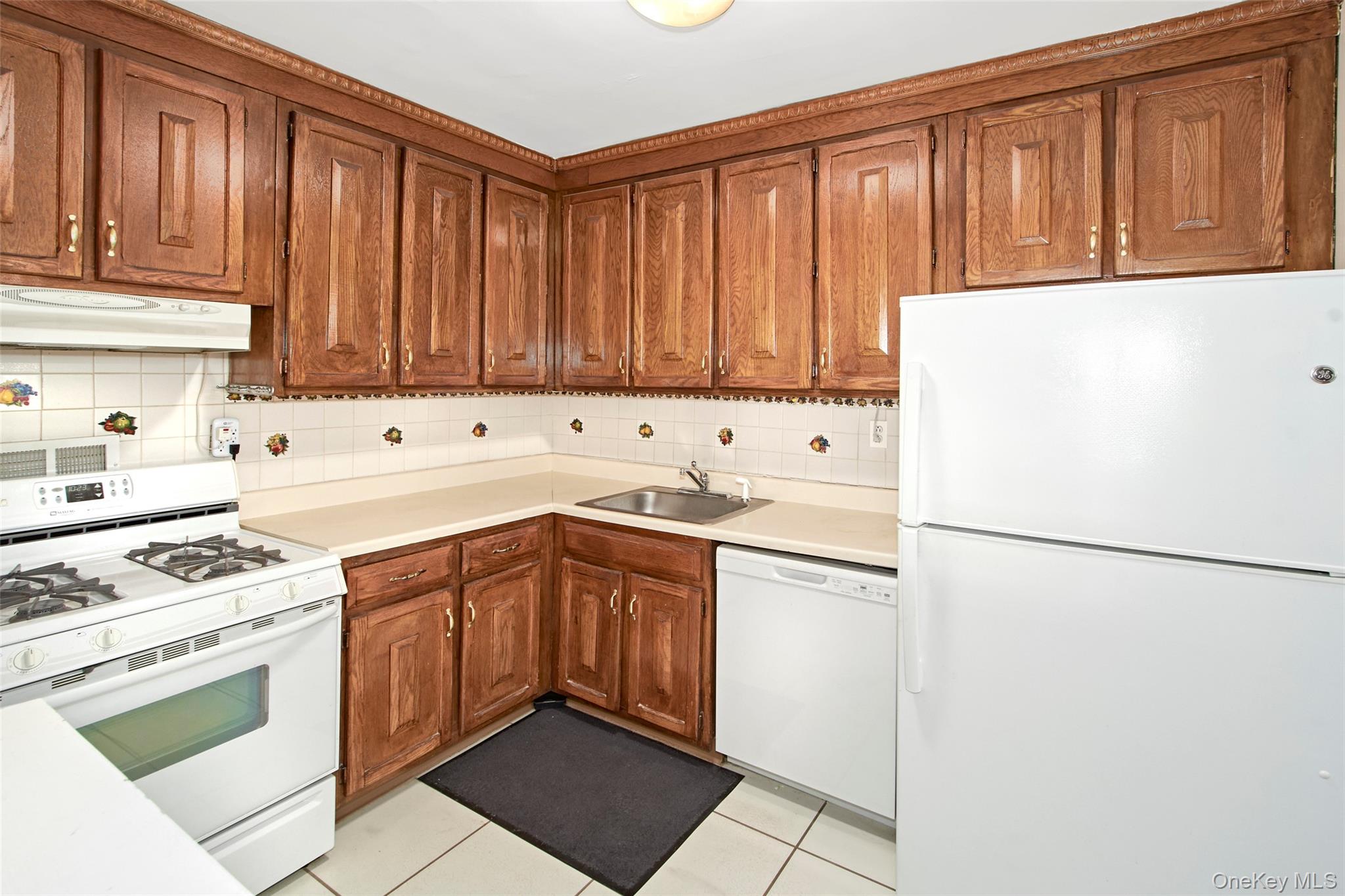 Kitchen with reddish-brown wood cabinets, cream counters, floral-accented tile backsplash, and white appliances.