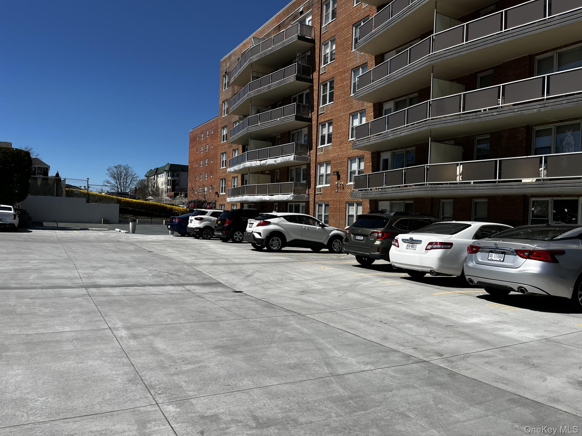 Red-brick apartment building with balconies beside a paved parking area lined with cars.