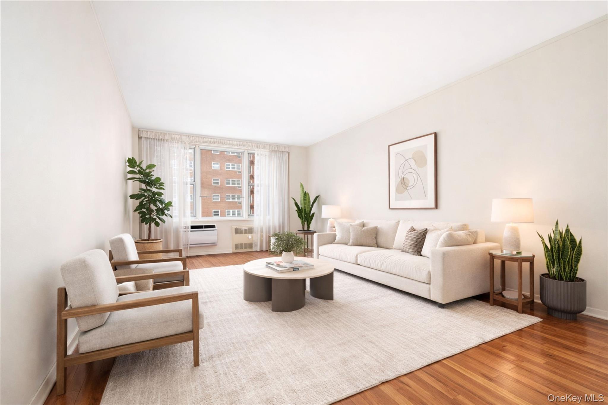Bright living room with a white sofa, wood-framed armchairs, plants, and large curtained windows.
