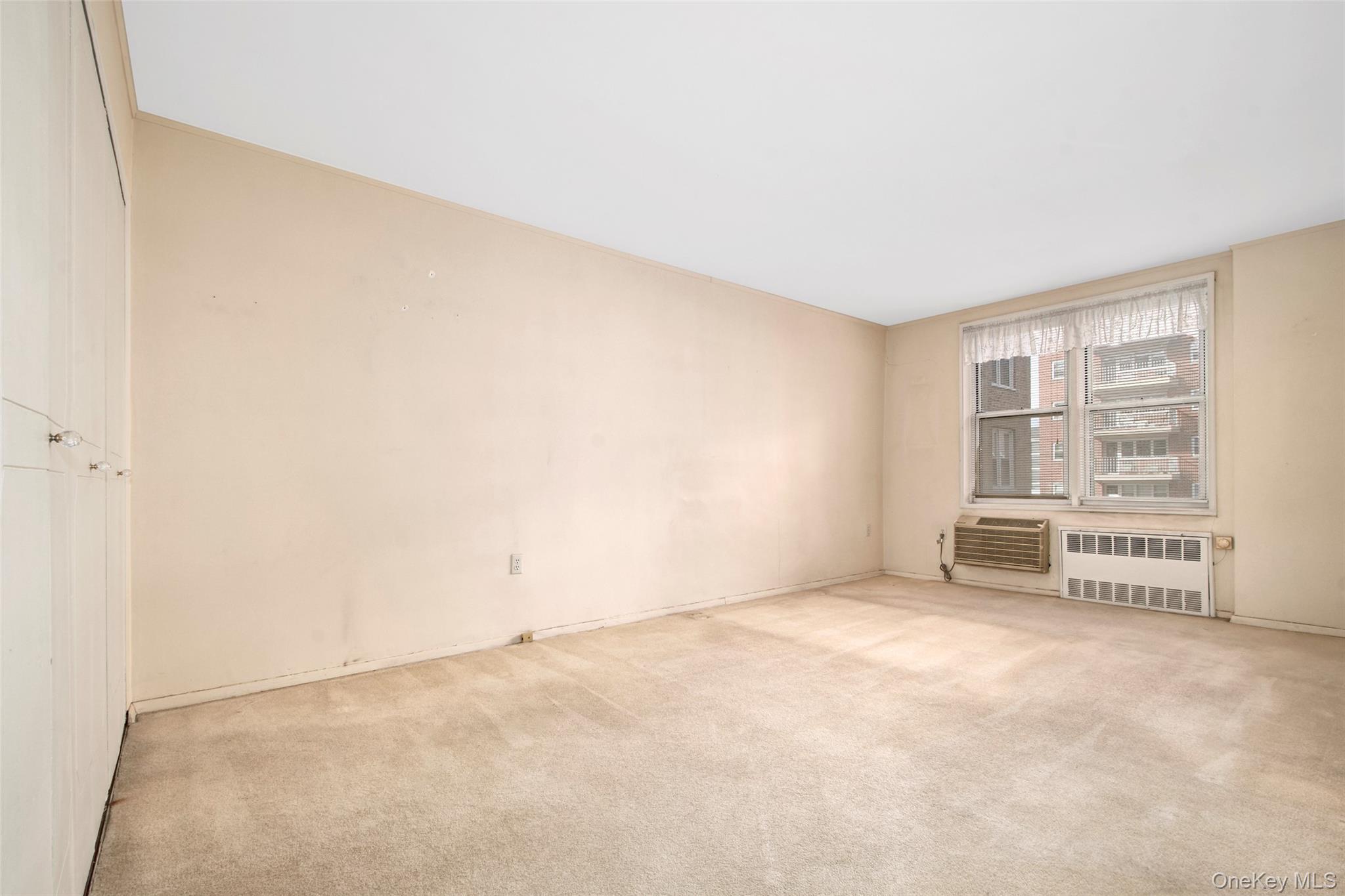 Empty carpeted bedroom with closet doors and a window above two air-conditioning units.