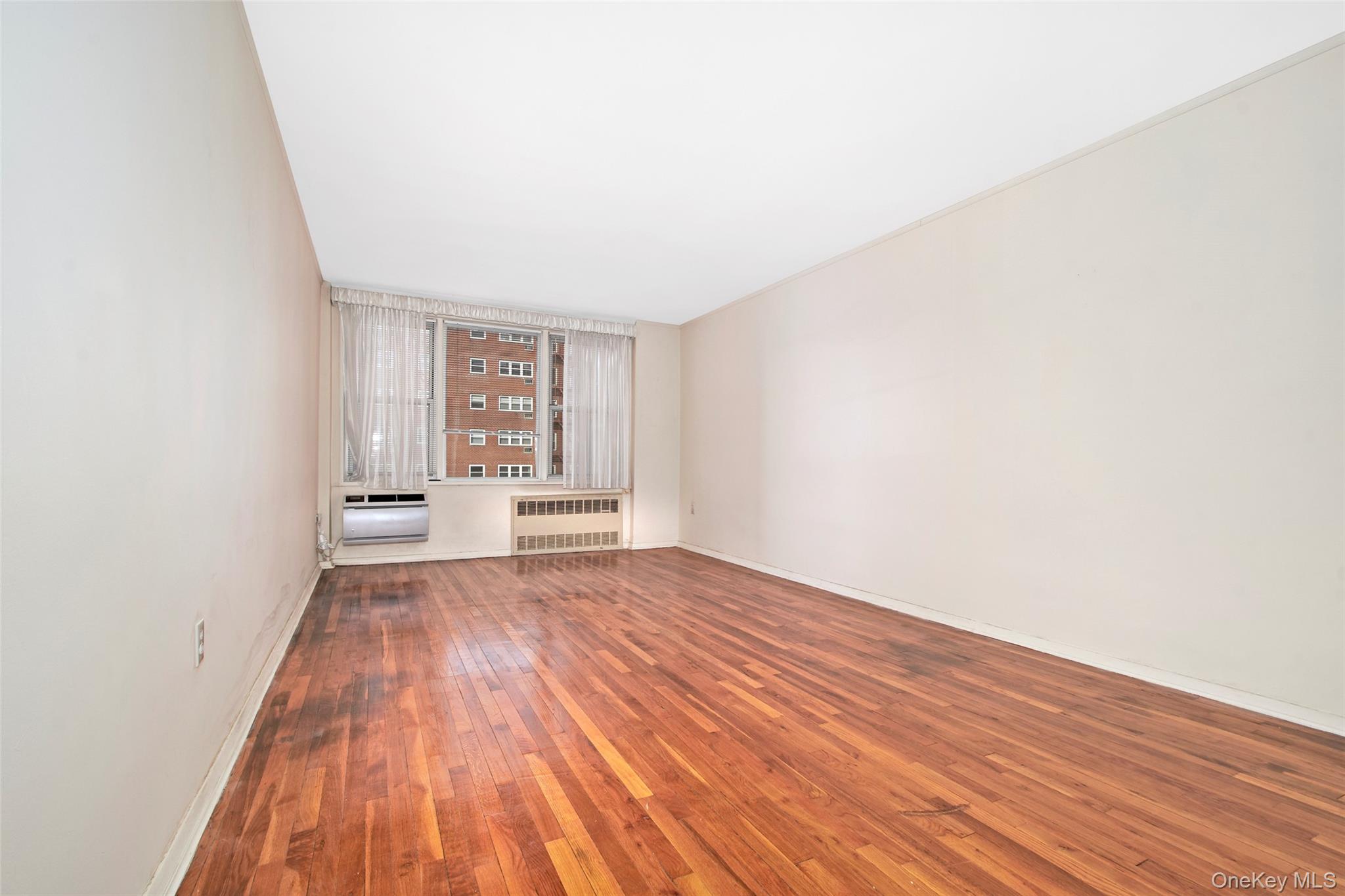 Empty living room with polished hardwood floors, a curtained window, and wall-mounted heating and air-conditioning units.