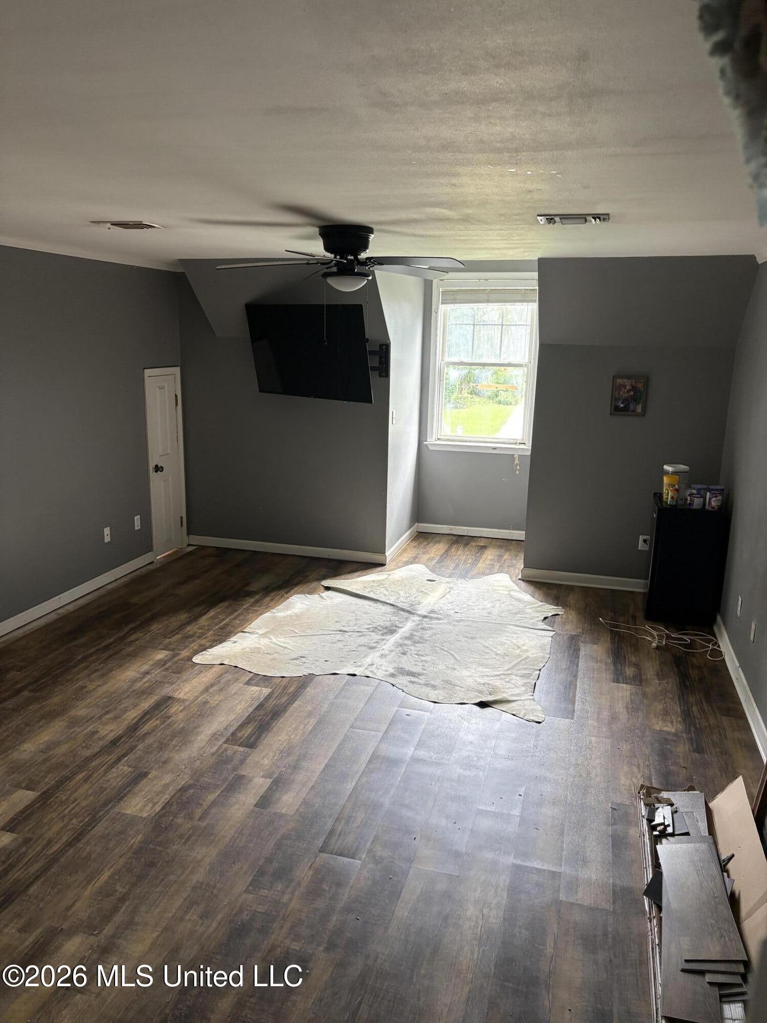 Sparse gray room with dark wood-look flooring, a ceiling fan, wall-mounted TV, and a bright window.
