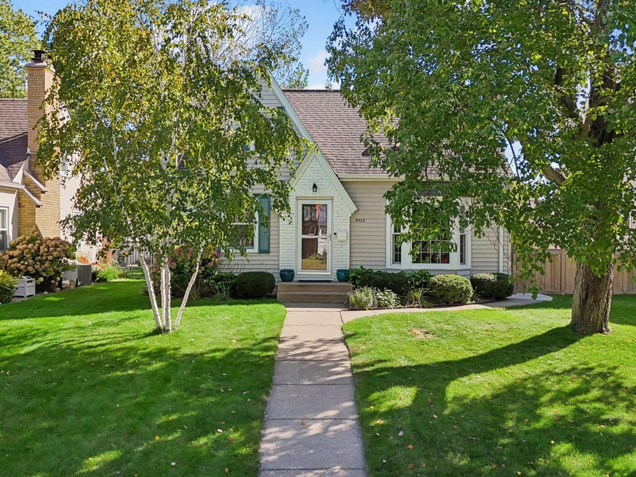 Light-colored gabled house behind mature trees, with a walkway crossing its green front lawn.