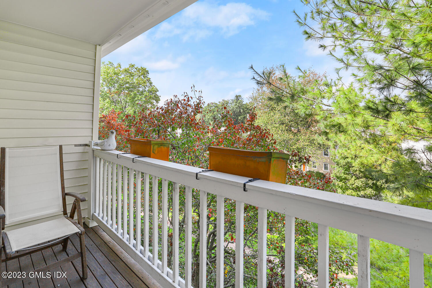 Apartment balcony with a white railing, chair, and planters overlooking autumn trees.