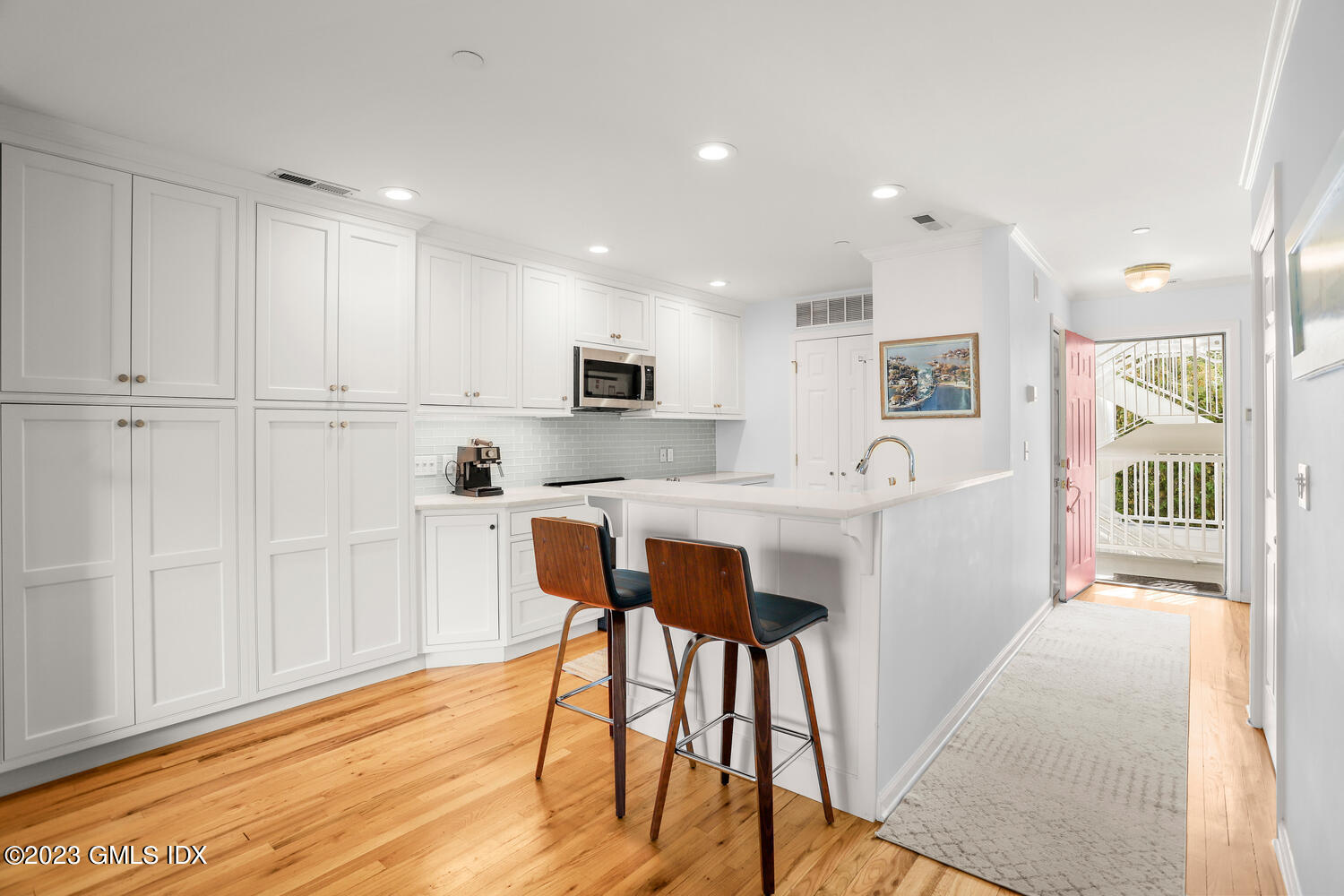 Bright white kitchen with a peninsula, two wooden bar stools, and an open pink front door.