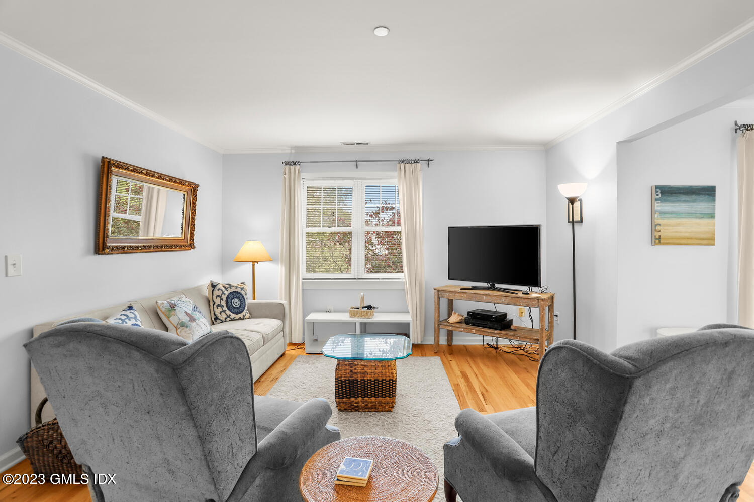 Living room with hardwood floors, a beige sofa, gray armchairs, and a television by a curtained window.