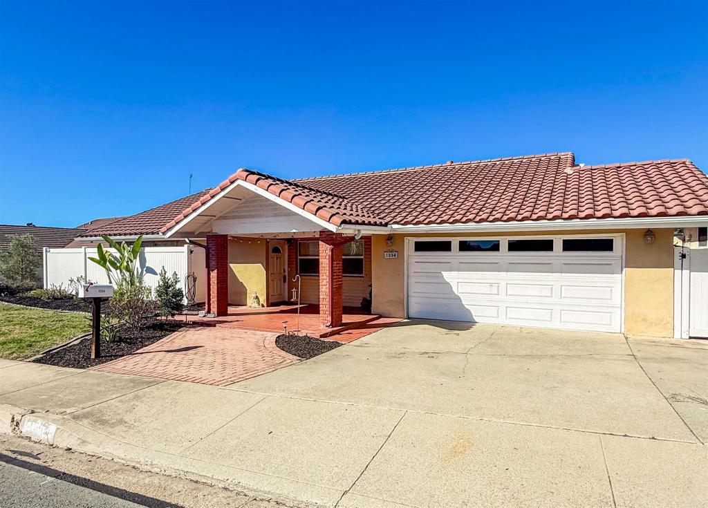 Single-story stucco house with a red tile roof, brick porch, and white double garage door.