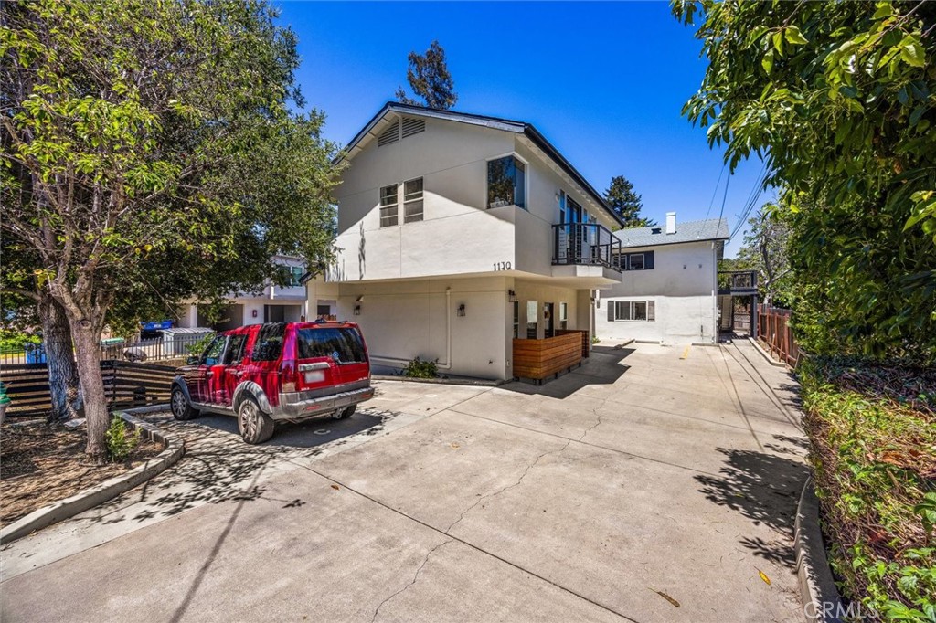 Two-story white building with an upper balcony and broad concrete driveway, framed by mature trees.