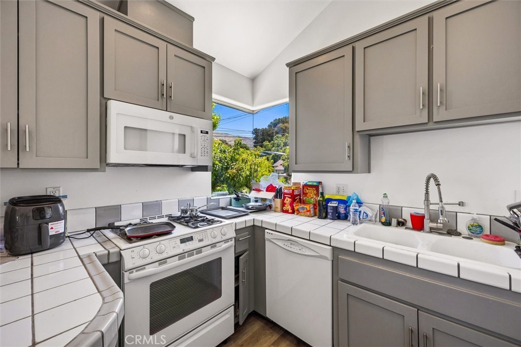 Kitchen with gray cabinets, white tile counters, and a window overlooking trees and a hillside.
