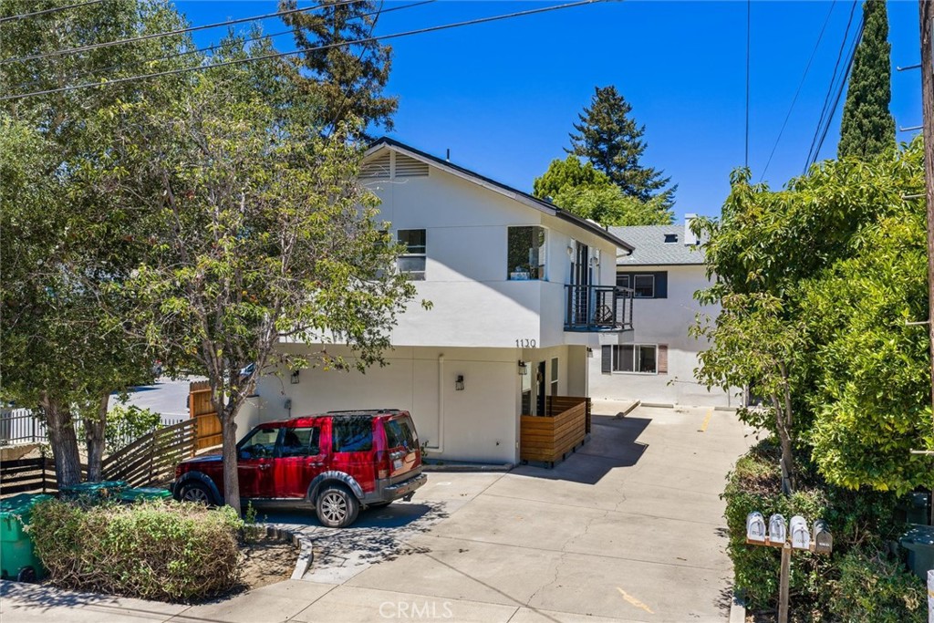 White two-story building with a small balcony, broad paved driveway, and red SUV parked in front.