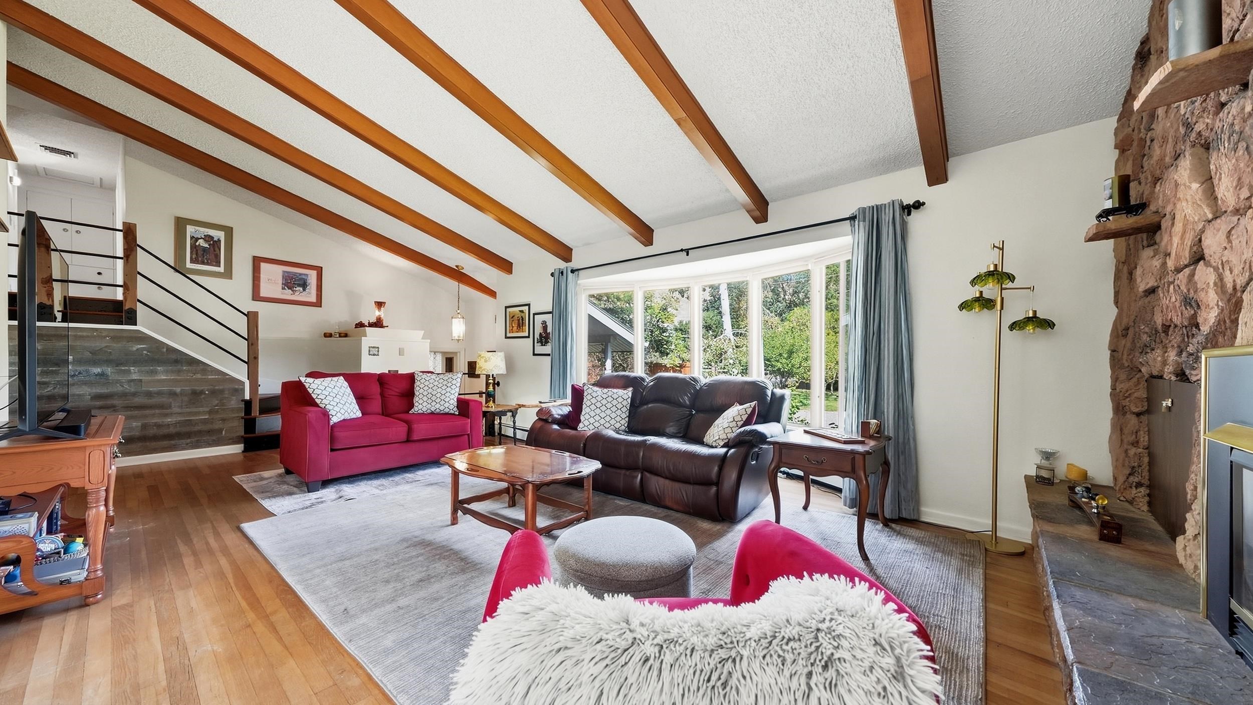 Living room with exposed wood beams, stone fireplace, and large windows overlooking greenery.