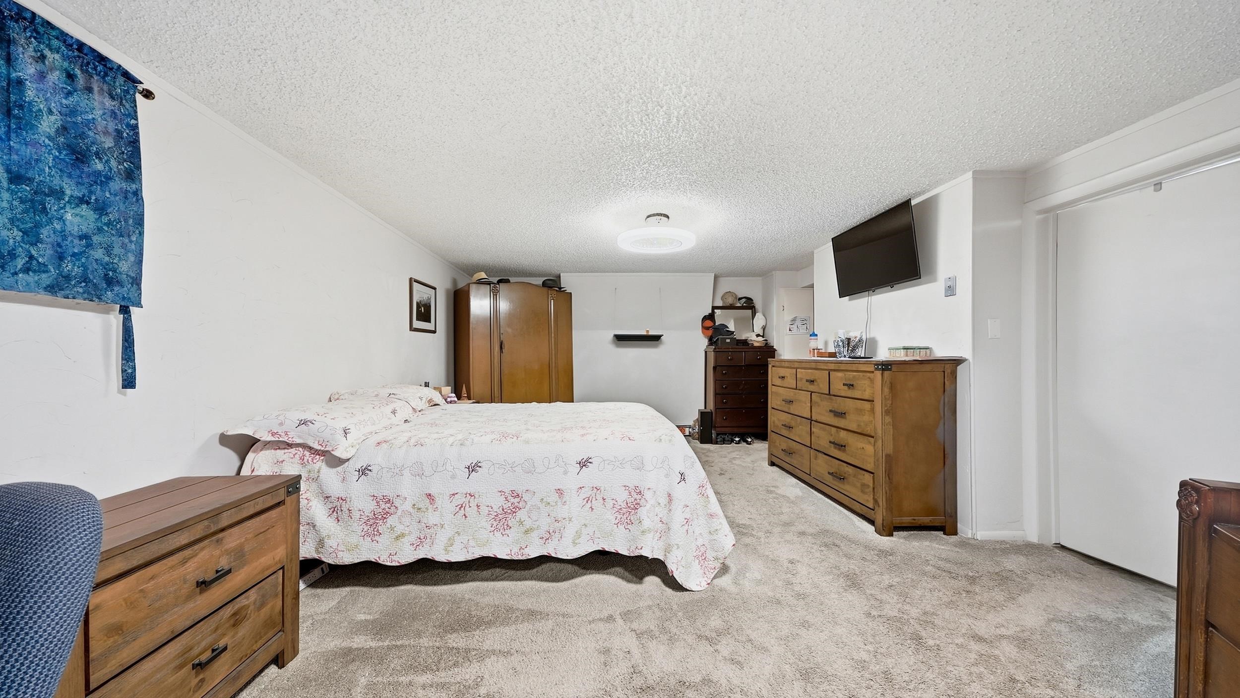 Bedroom with a floral quilt, wood dressers, beige carpet, and wall-mounted television.