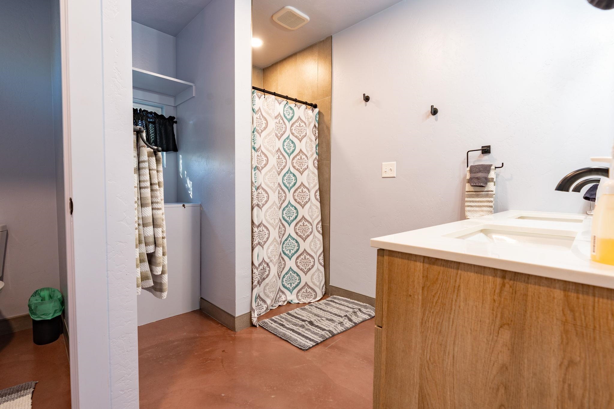 Bathroom with wood-front vanity, tiled shower, patterned curtain, and terracotta-colored floor.