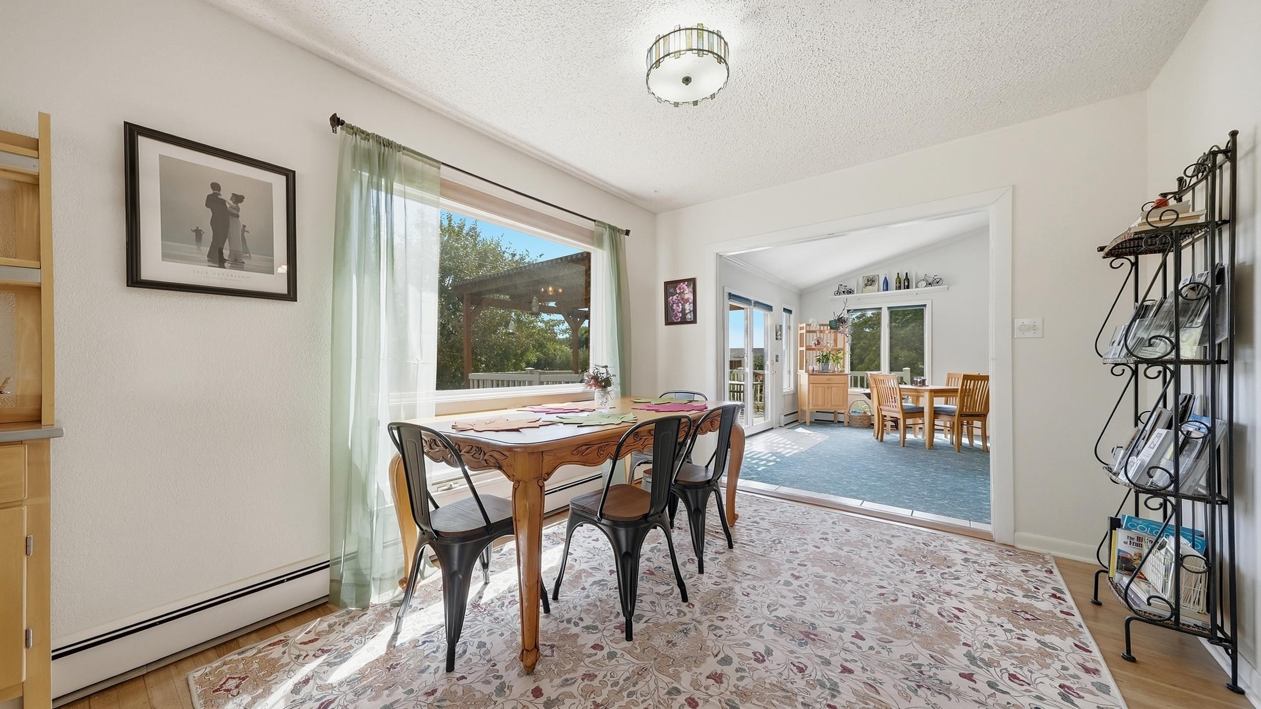 Dining room with a carved wooden table, black chairs, floral rug, and a wide opening to a bright adjoining room.