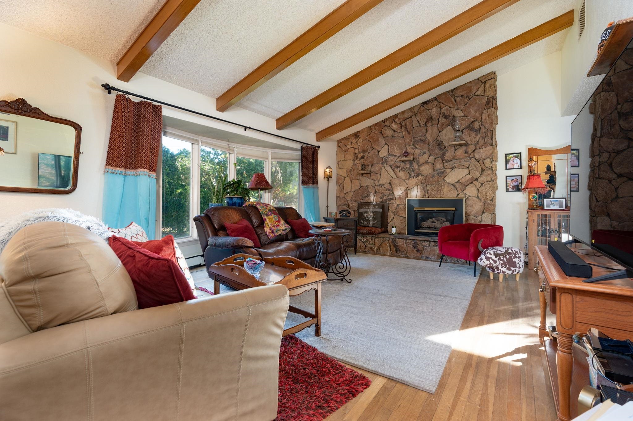 Living room with exposed ceiling beams, a stone fireplace, and large windows.