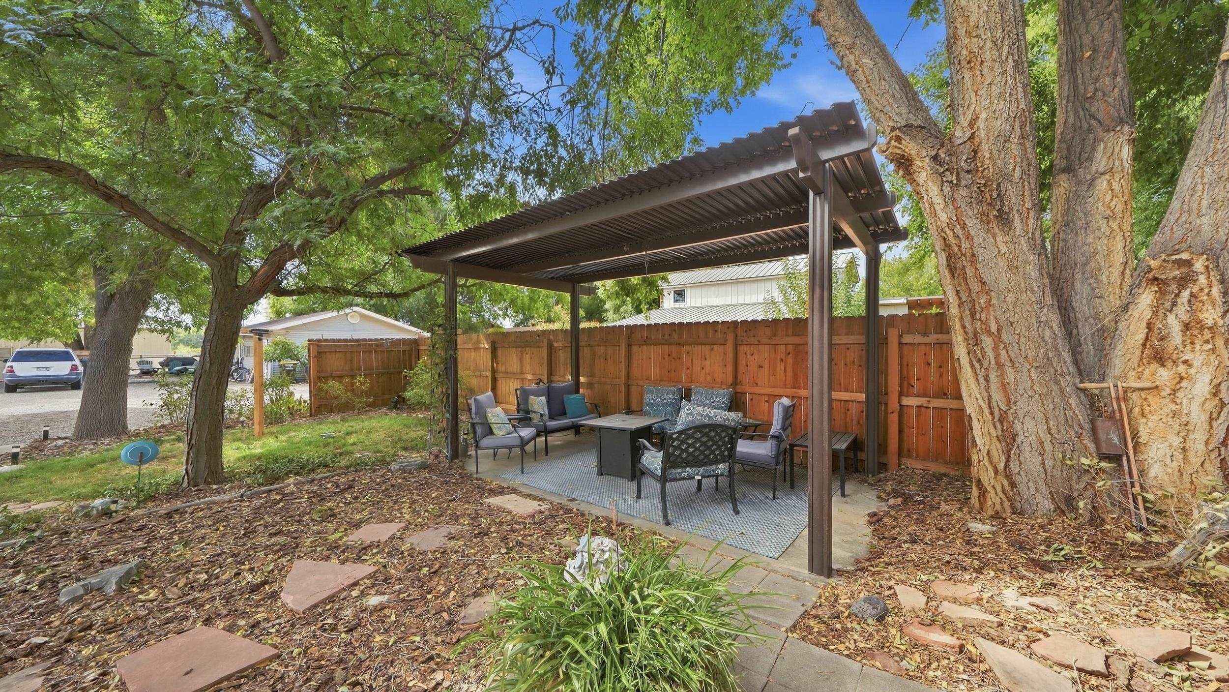 Furnished backyard patio under a pergola, shaded by mature trees and enclosed by a wooden fence.