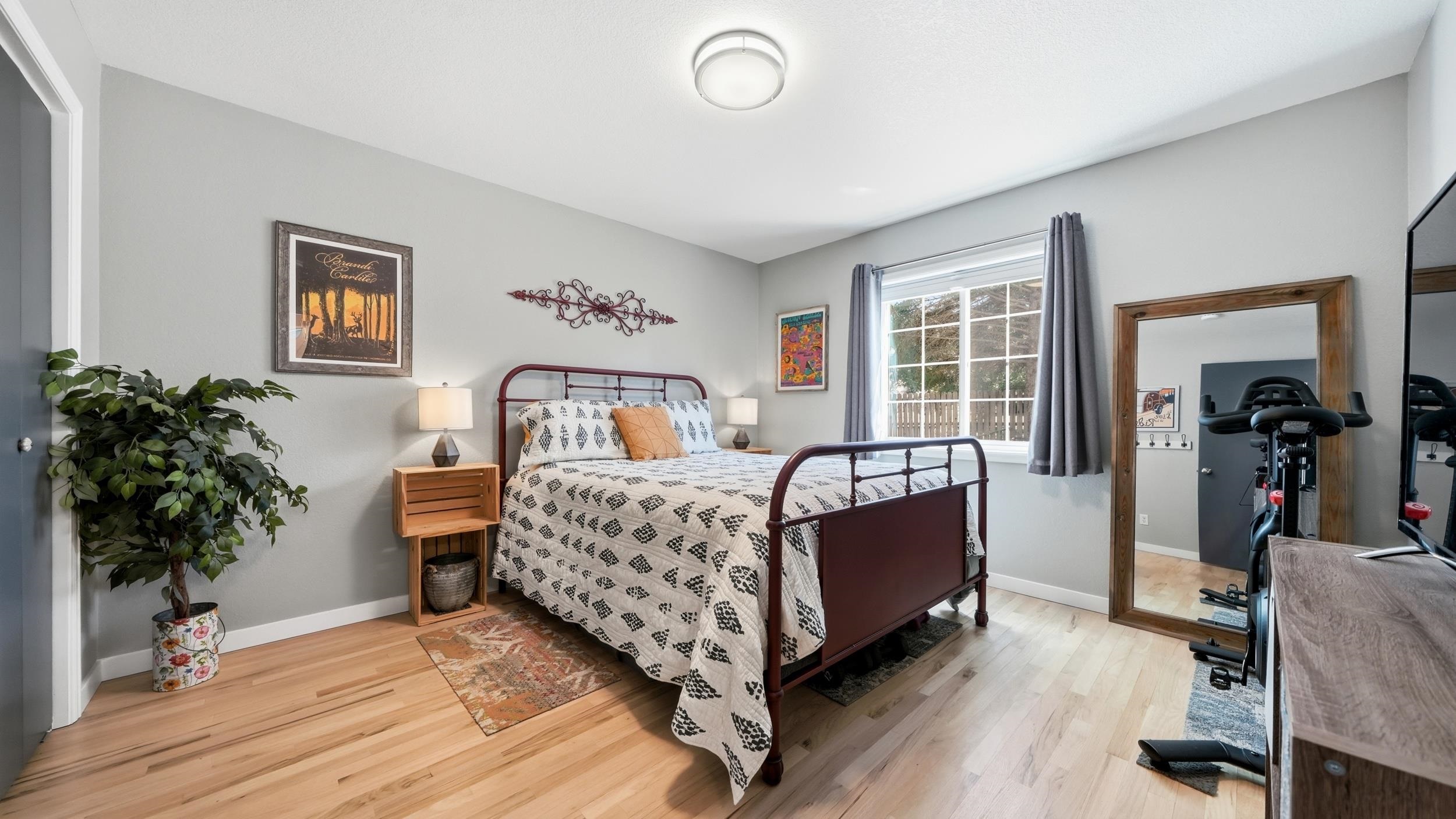 Furnished bedroom with a dark metal bed, patterned bedding, and a wood-framed mirror reflecting exercise equipment.