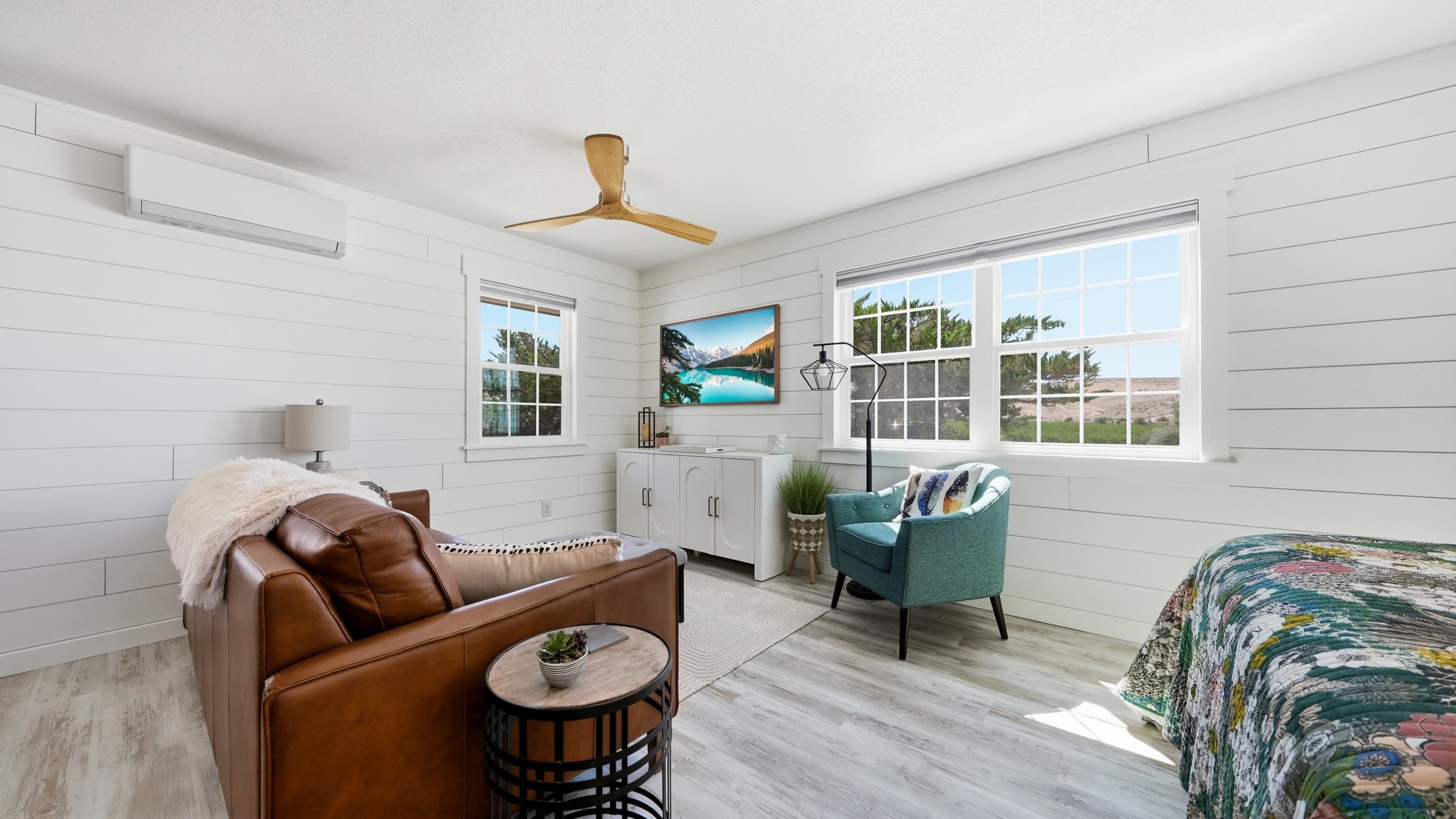 Staged bedroom with a brown leather sofa, teal armchair, and large windows overlooking open terrain.