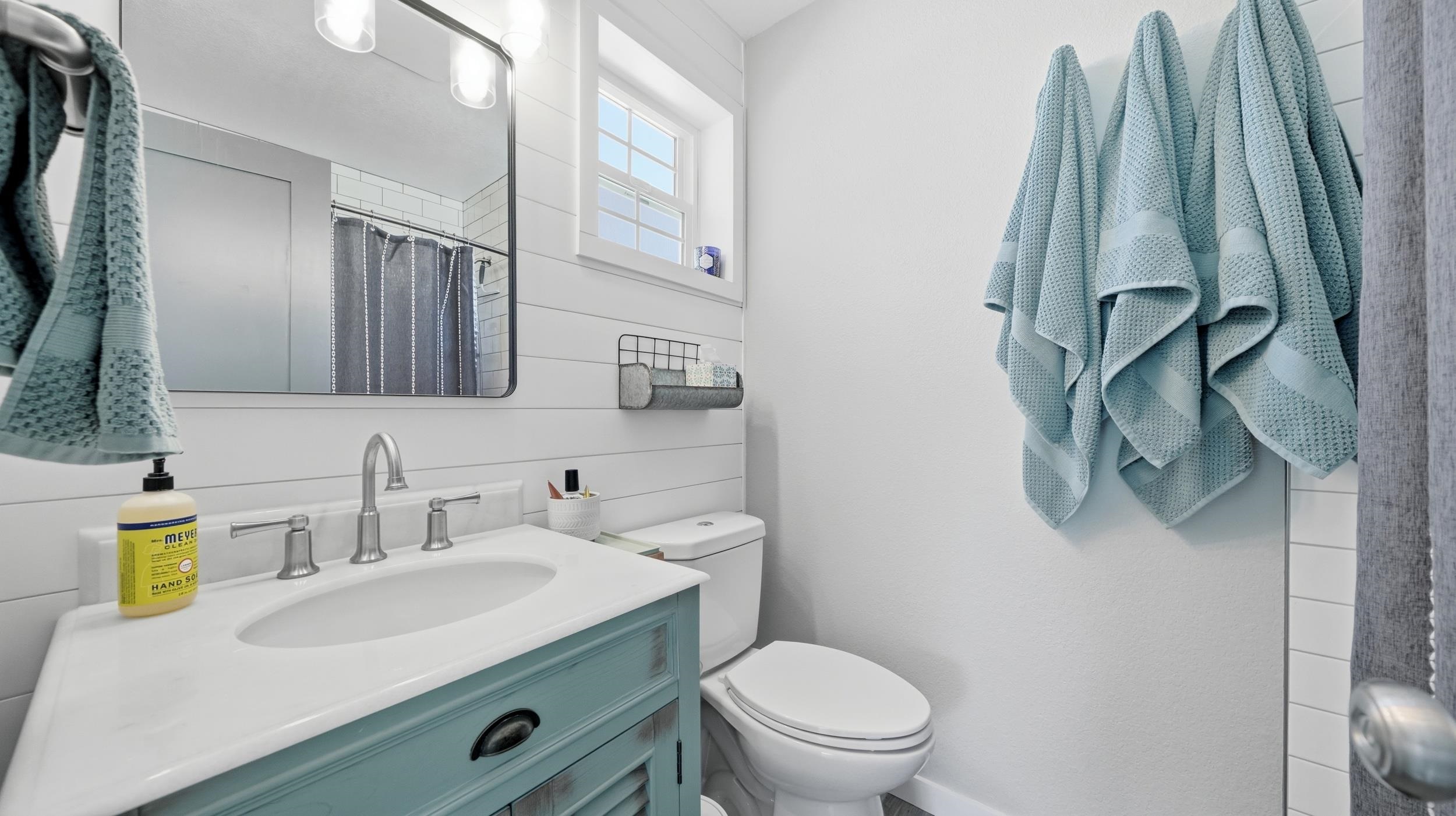 Bathroom with a pale blue-green vanity, white shiplap walls, high window, and hanging blue towels.