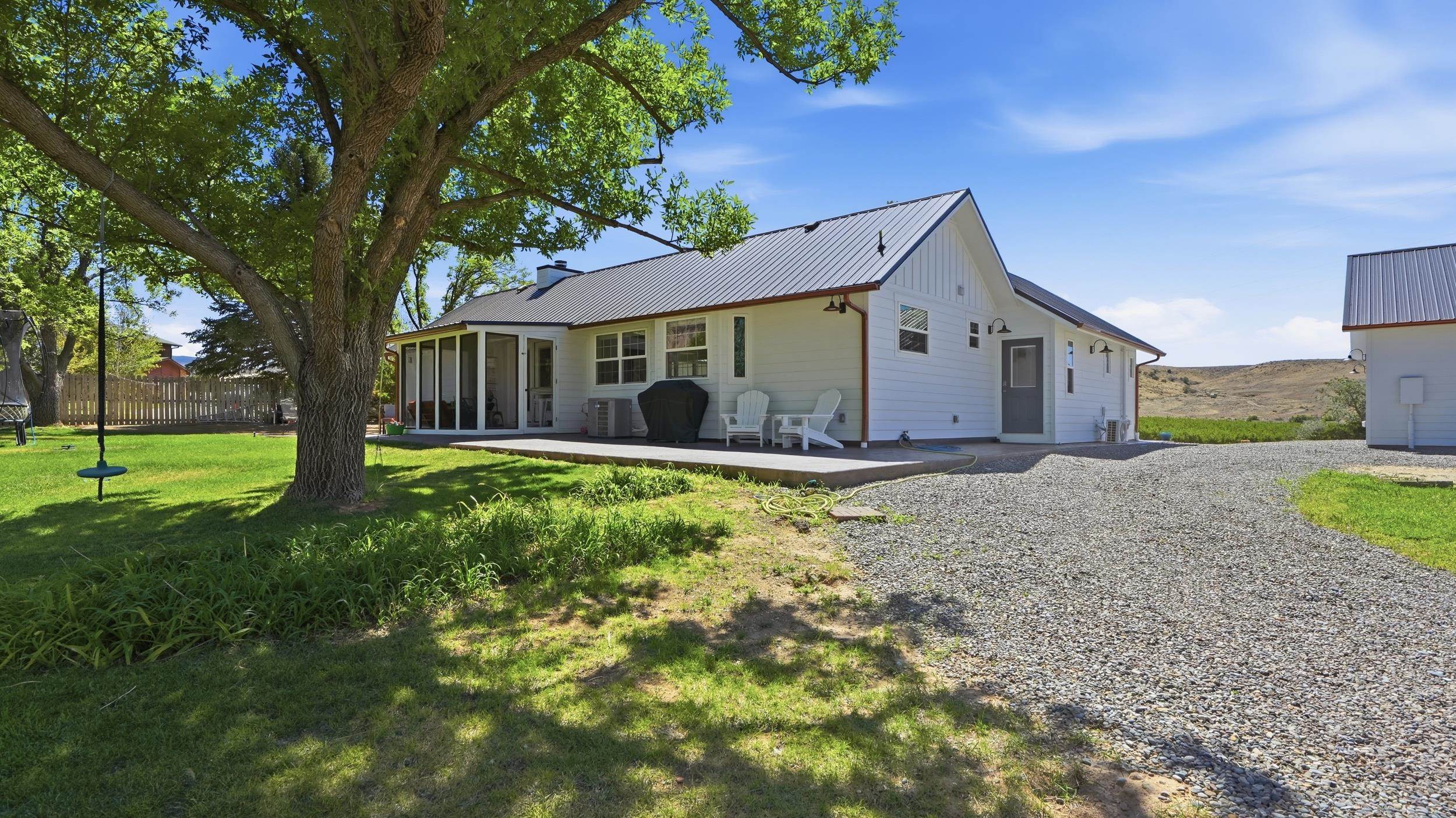 White home with a metal roof, screened porch, patio chairs, large shade tree, and gravel driveway.