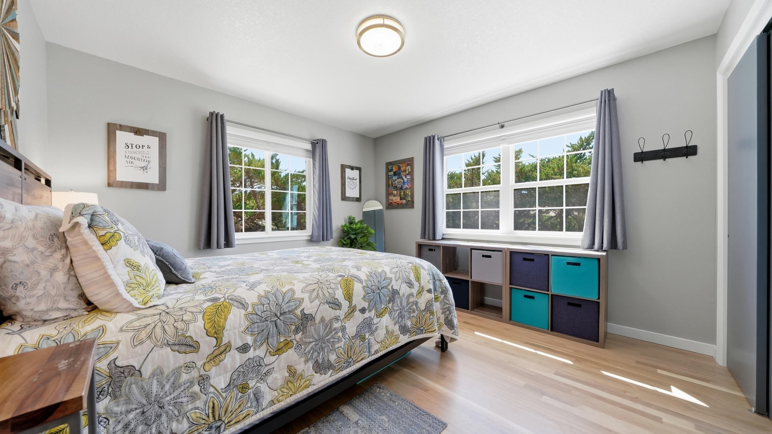 Furnished bedroom with a floral bedspread, two large windows, and colorful storage bins.