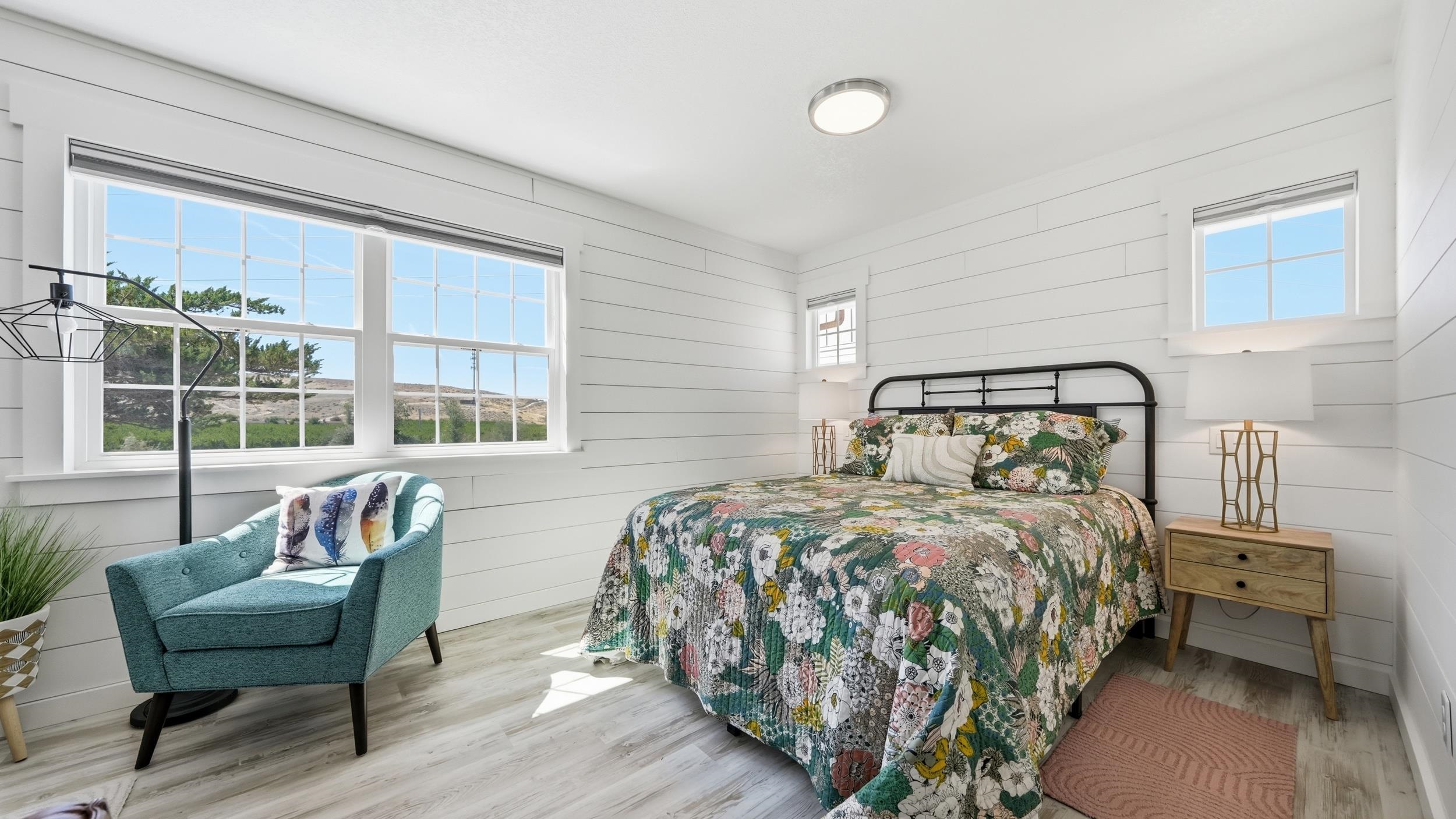 Bedroom with a floral quilt, teal chair, and large windows overlooking rolling hills.