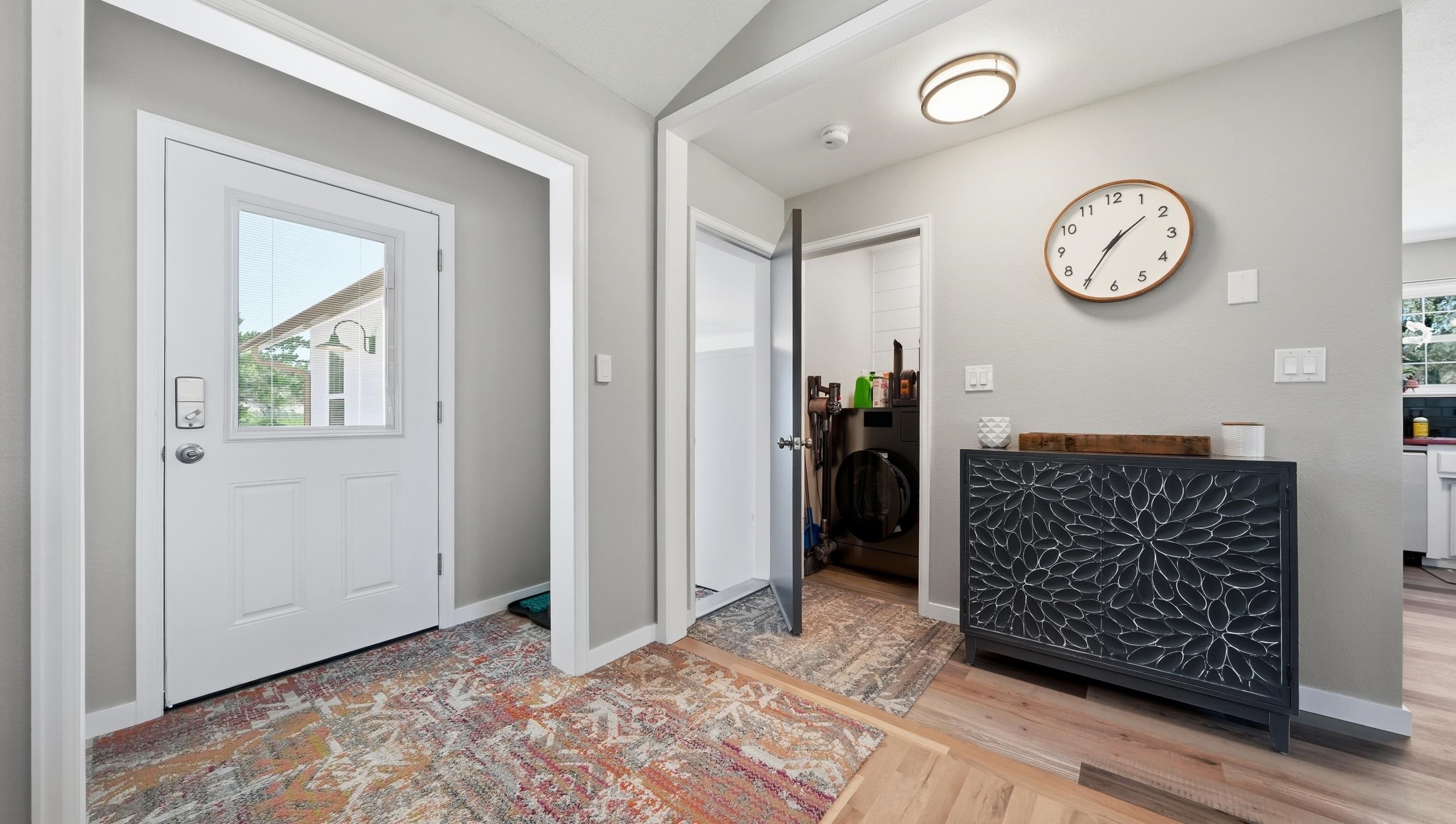 Entryway with a white front door, patterned rug, black console cabinet, and laundry appliances behind an open door.