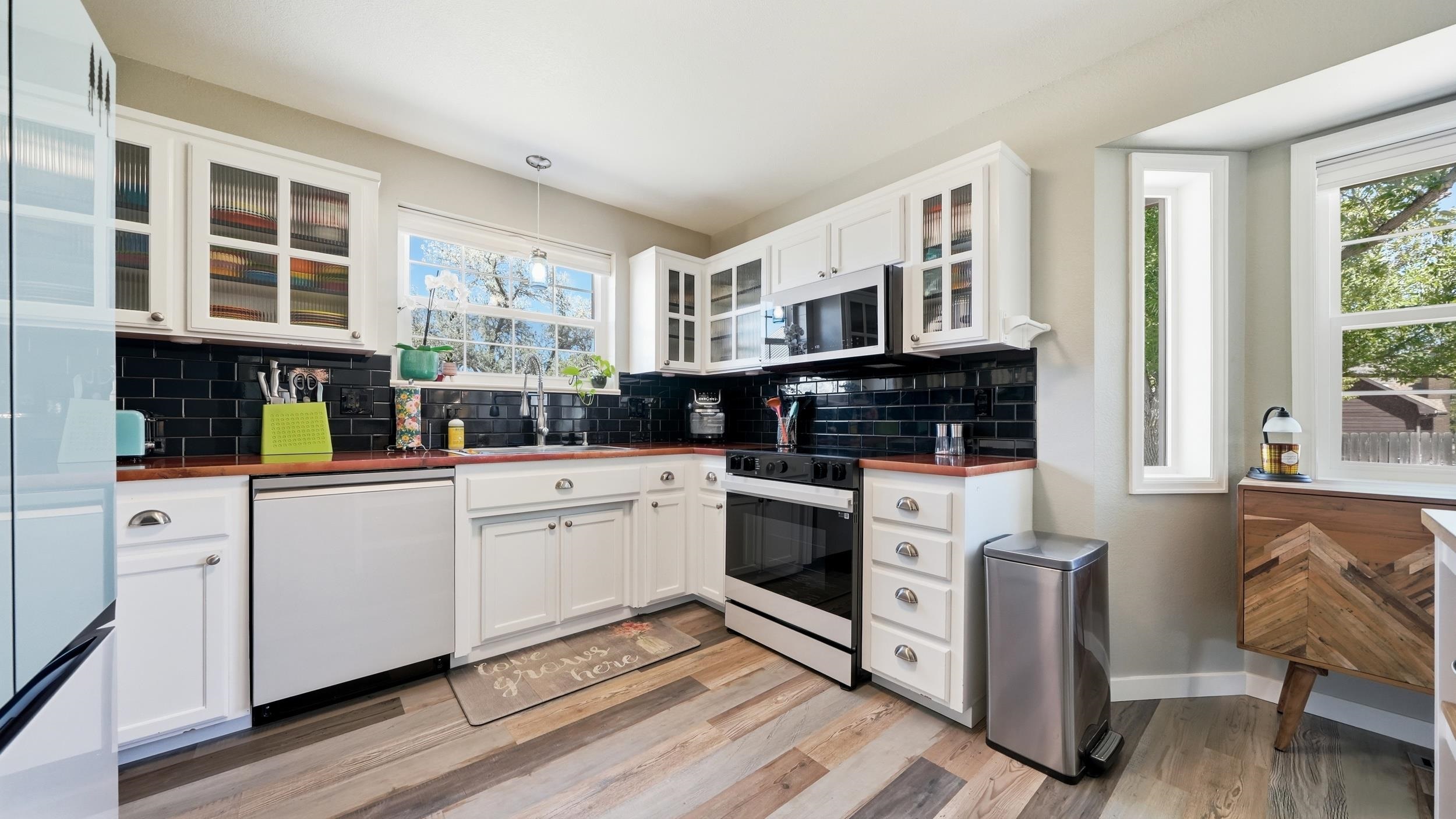 Bright kitchen with white cabinets, black tile backsplash, reddish countertops, and stainless-steel appliances.