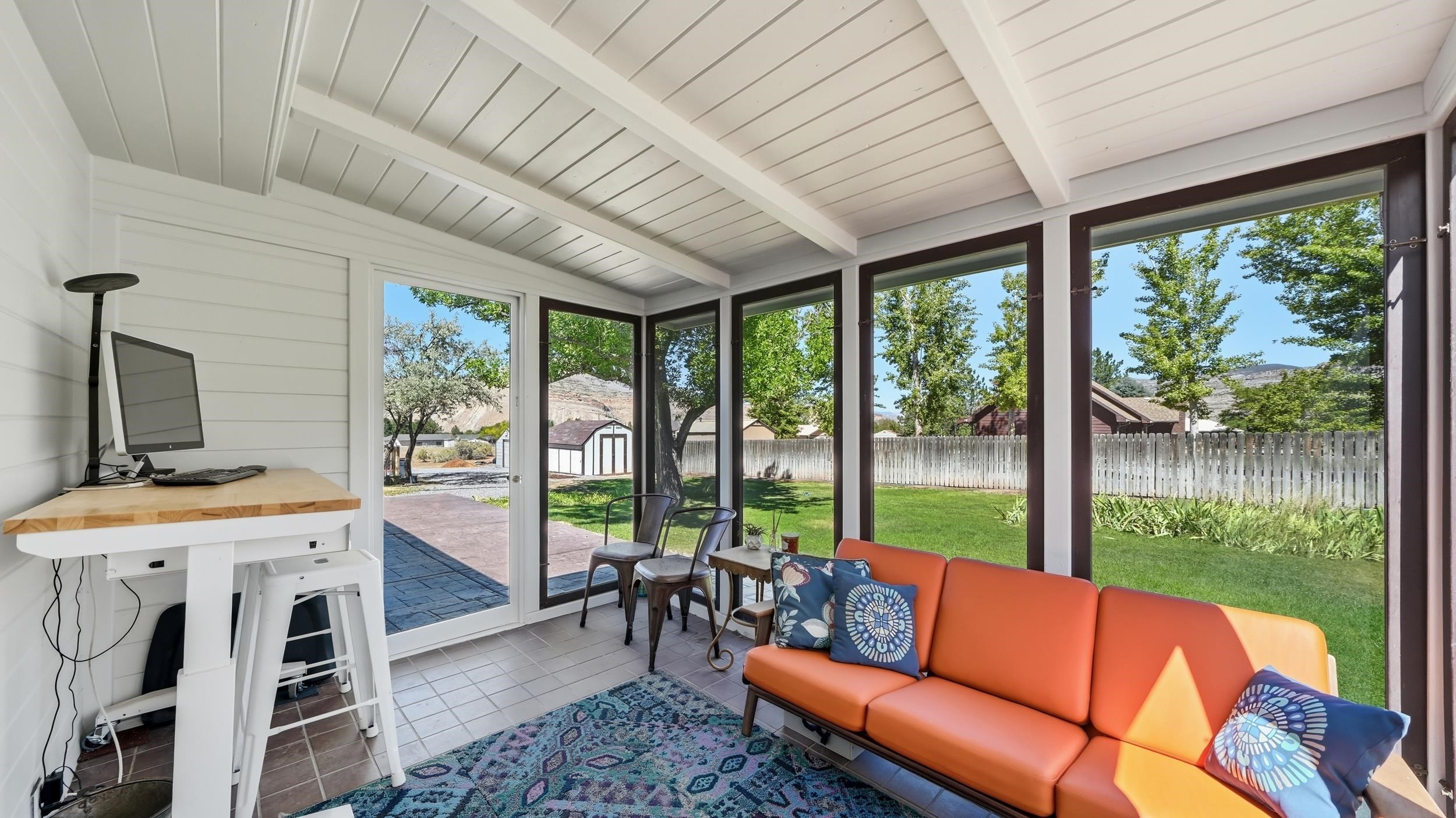 Enclosed sunroom with an orange sofa, desk, and windows overlooking a fenced yard and rocky hills.