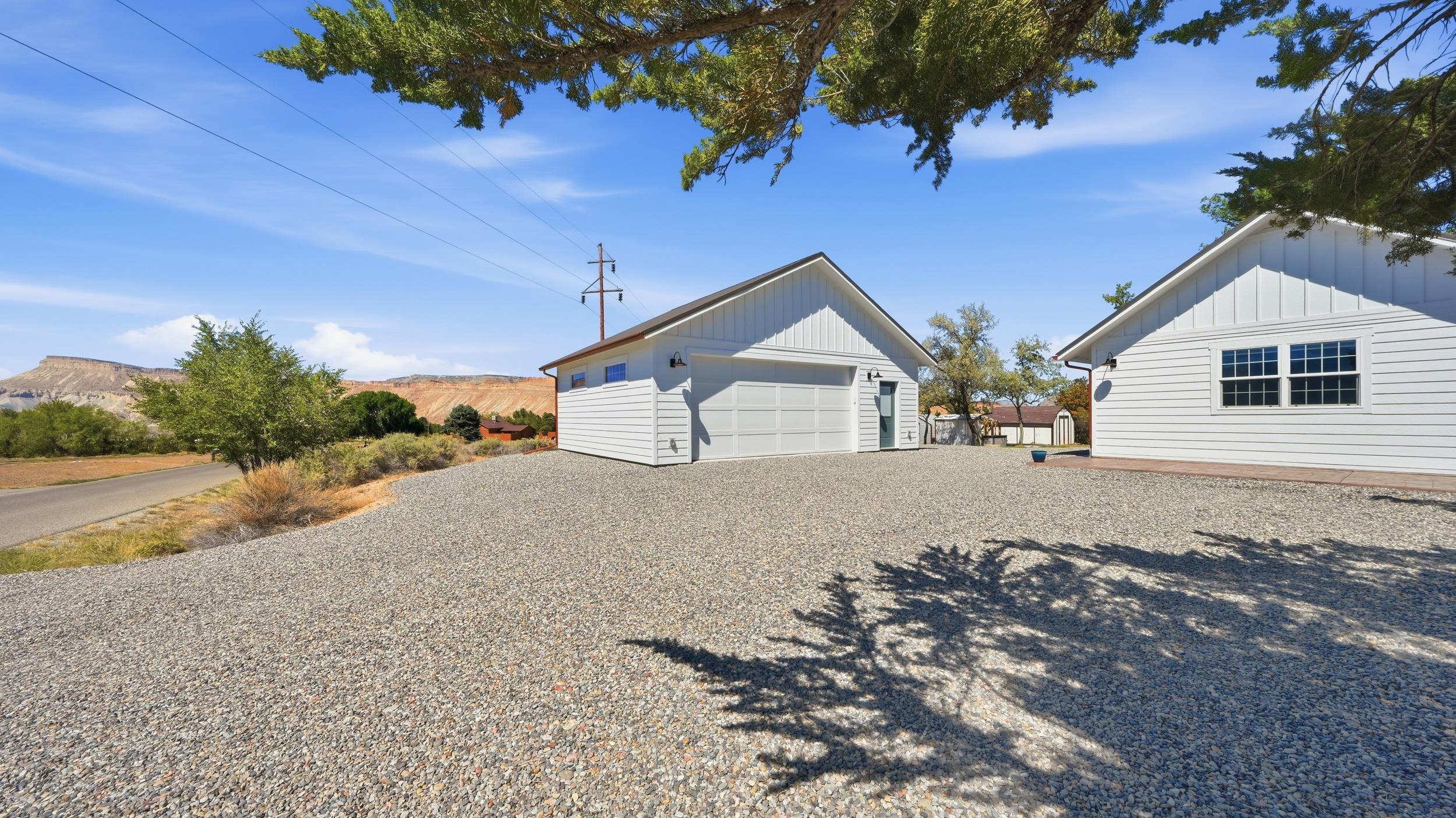 White home and detached garage beside a broad gravel driveway, with mesas in the distance.