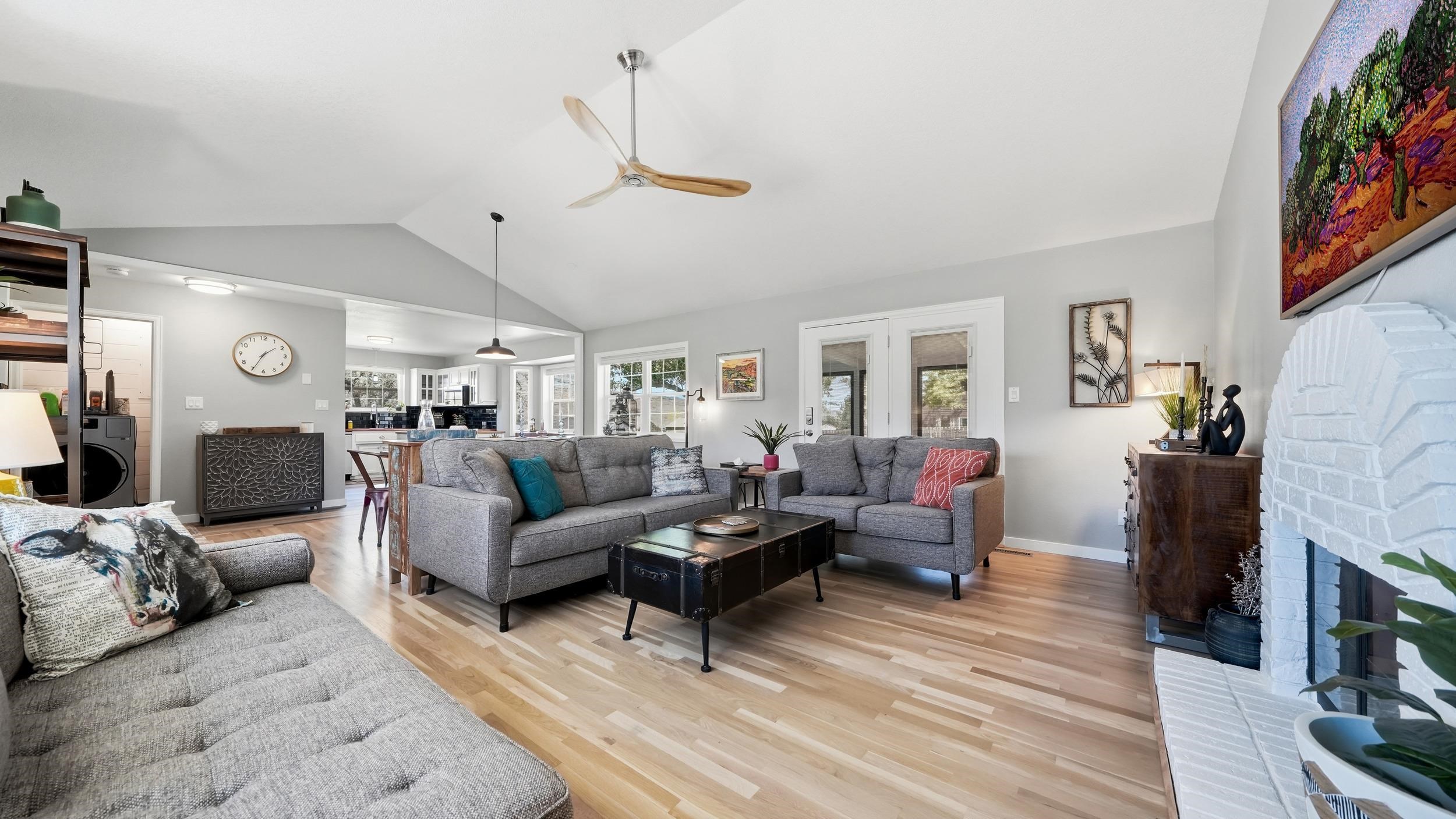 Living room with gray sofas, vaulted ceiling, light wood floors, and a white brick fireplace.