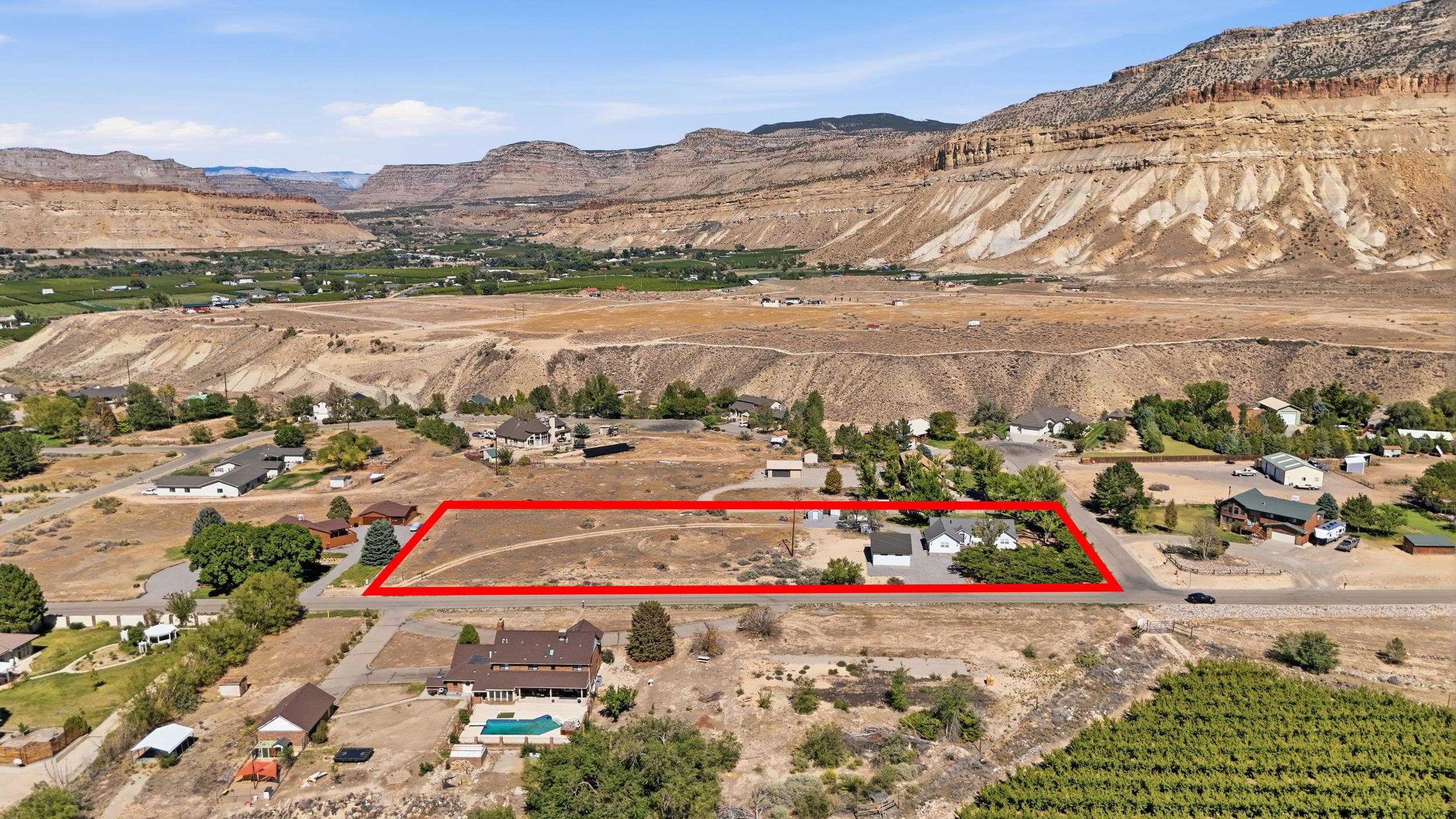 Aerial view of a red-outlined parcel beside a road, with homes and rocky cliffs in the distance.
