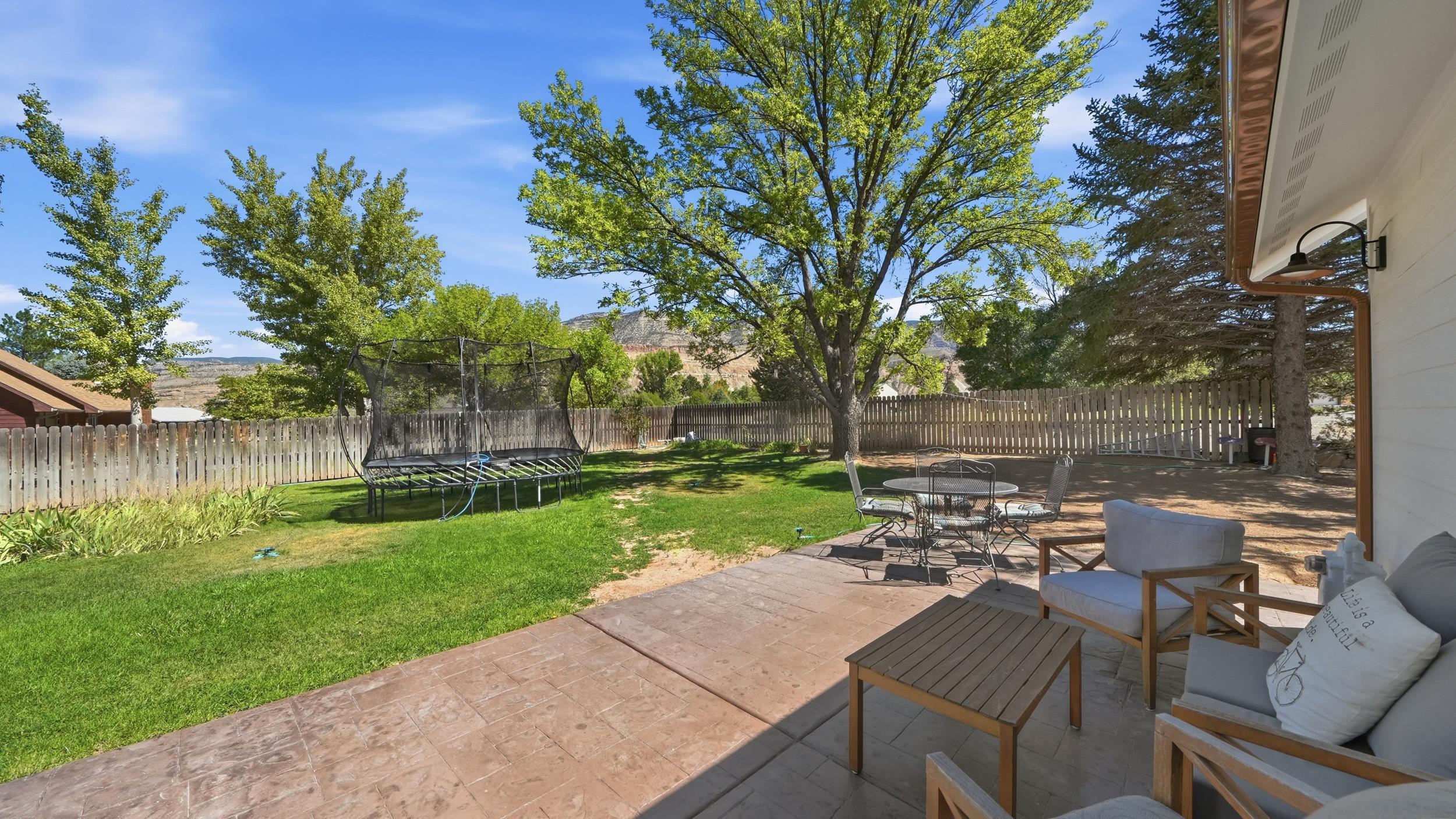 Backyard patio with outdoor seating overlooking a fenced lawn, trampoline, mature trees, and distant hills.