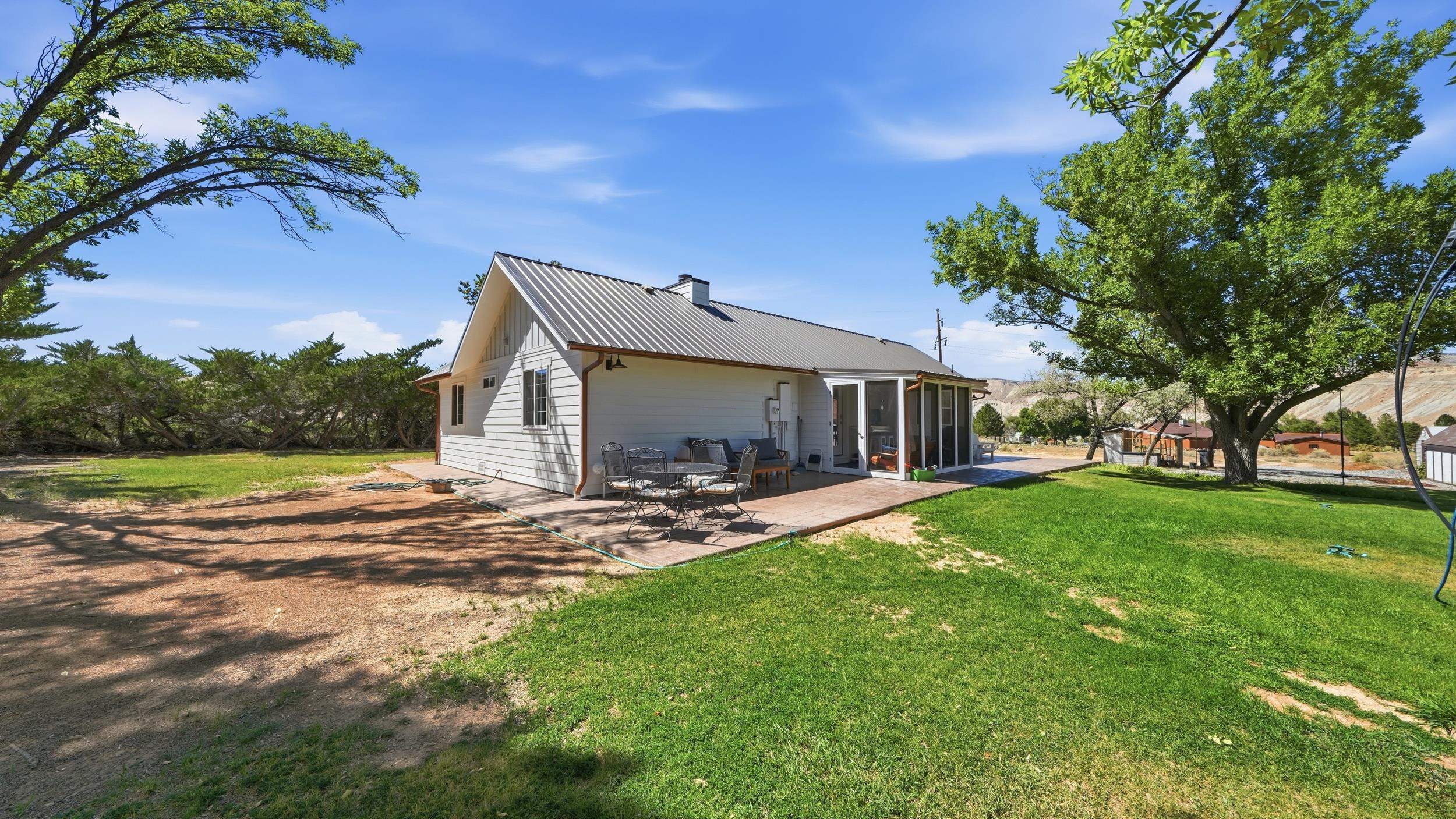 House rear with a metal roof, patio seating, lawn, and mature trees.