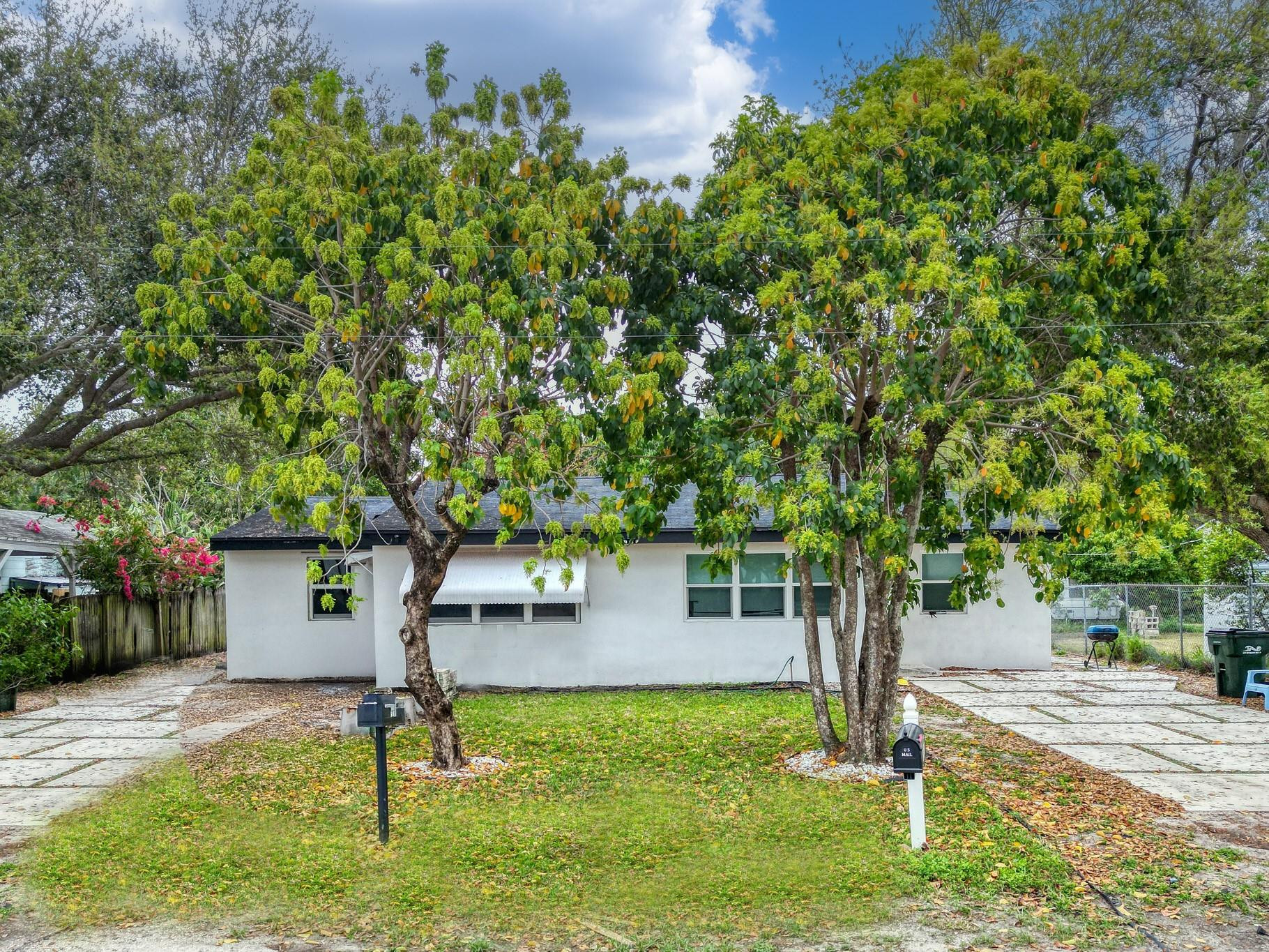 Single-story white house shaded by mature trees, with a front window awning and grassy yard.