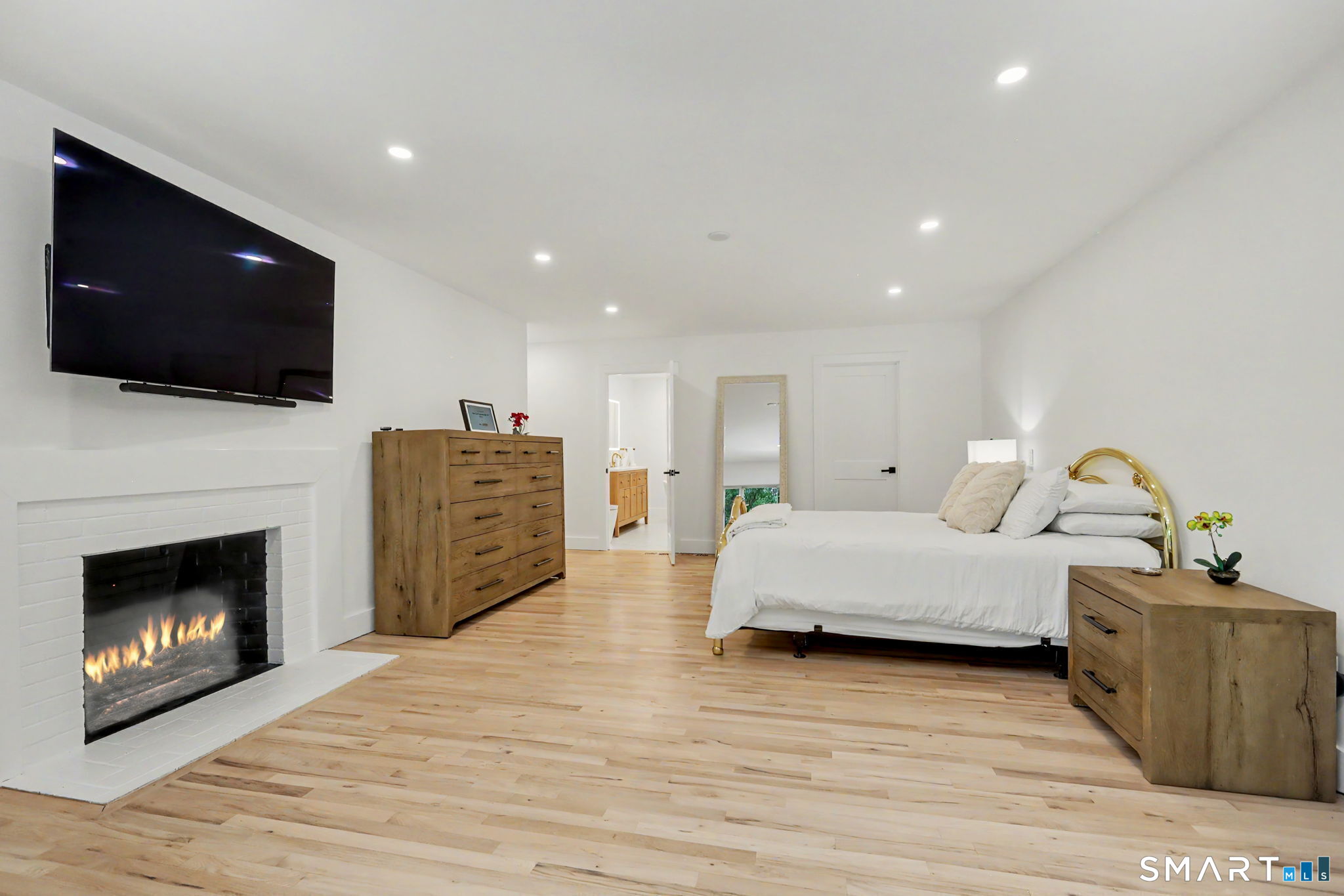 Bedroom with a gold headboard, light wood furnishings, wall-mounted TV, and lit fireplace.