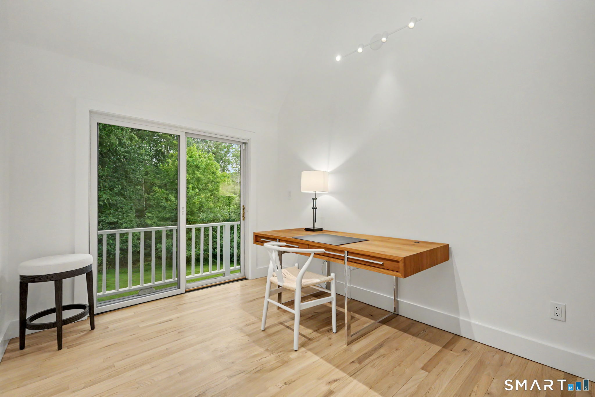 Bright home office with a wooden desk beside sliding glass doors overlooking leafy trees.
